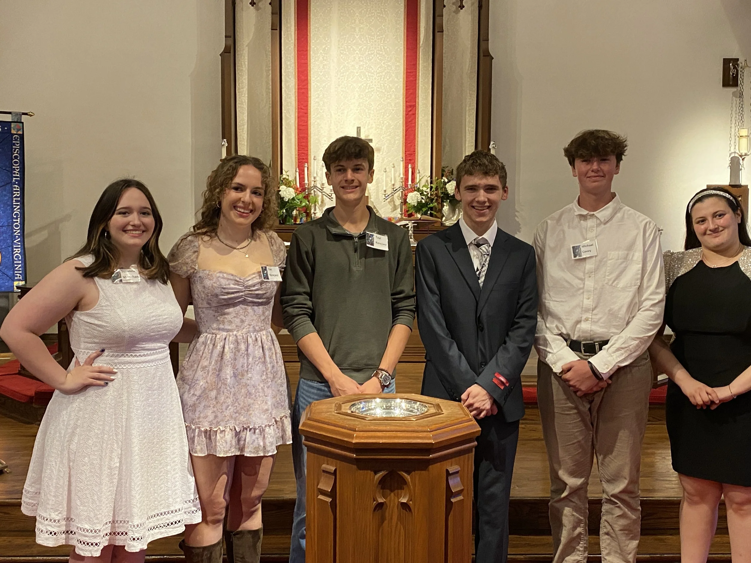 Group of six young people standing inside a church, dressed in formal and semi-formal attire, smiling at the camera with an altar and floral arrangements in the background.