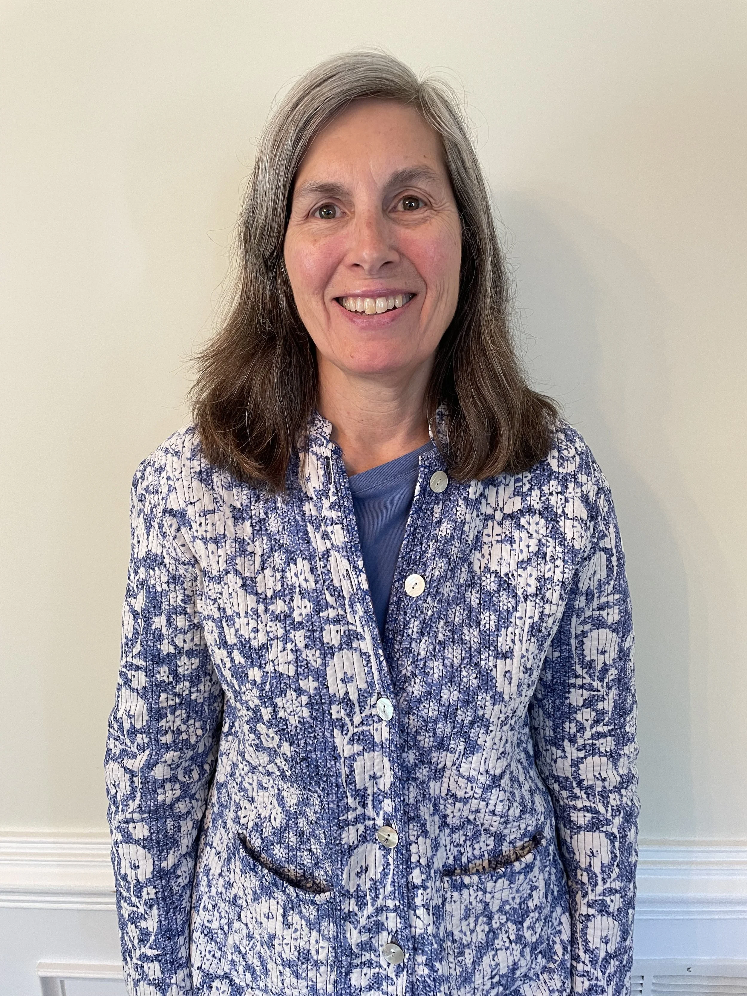 A middle-aged woman with shoulder-length brown hair and a warm smile, wearing a blue patterned jacket with white buttons and a blue shirt underneath, standing against a plain light-colored background.