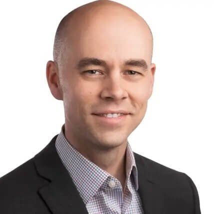 Professional portrait of a bald man in a suit and checkered shirt smiling against a white background.