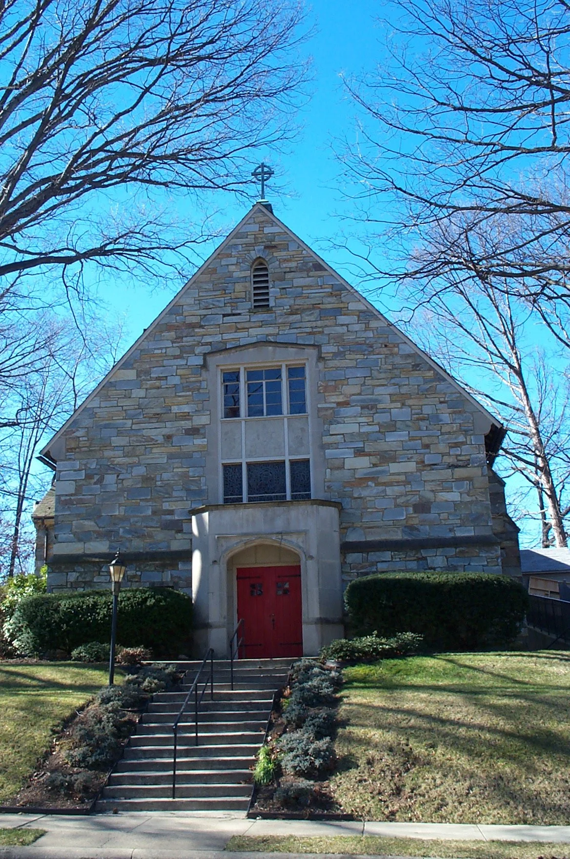 A stone church with a red door, set between bushes and trees, with a flight of steps leading to the entrance, under a clear blue sky.