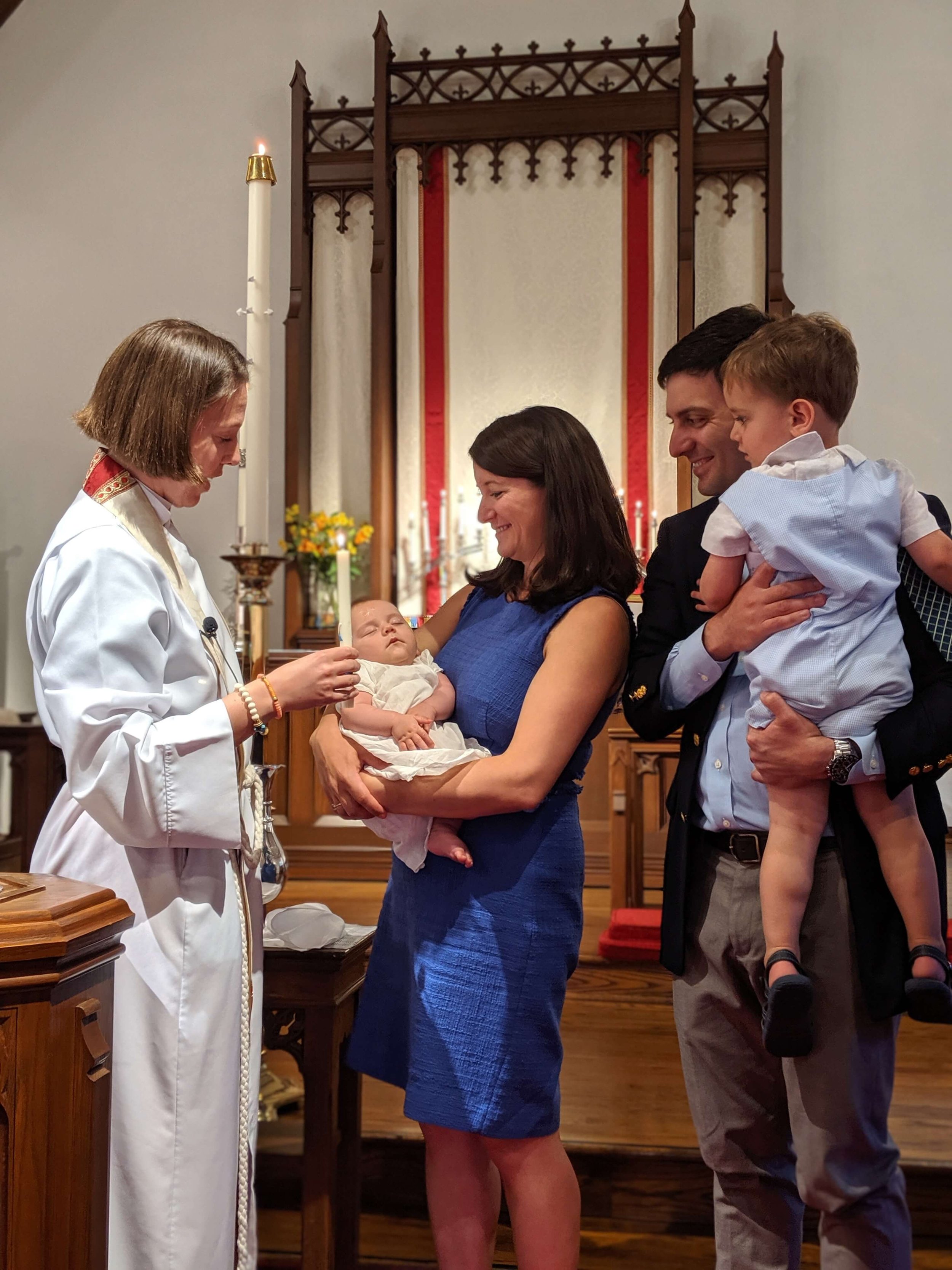 A family participating in a baptism ceremony in a church. A woman holds a baby while a priest administers the baptism. A man and child stand nearby, observing the event.