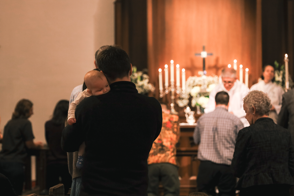 A man holding a small child during a church service or baptism ceremony, with an altar and congregation in the background.