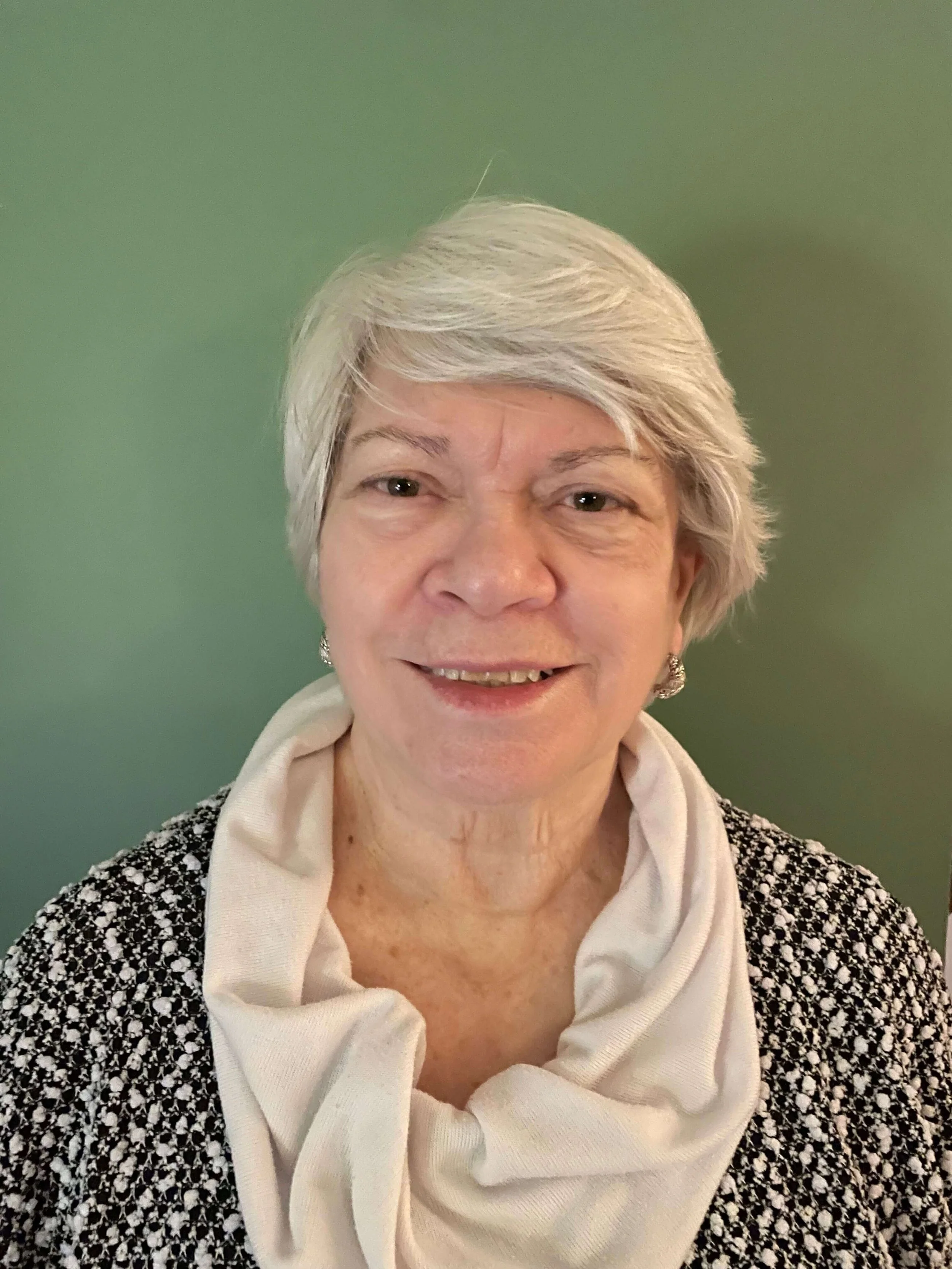 An elderly woman with short gray hair smiling, wearing a patterned jacket and a light-colored cowl neck top, with a green background.