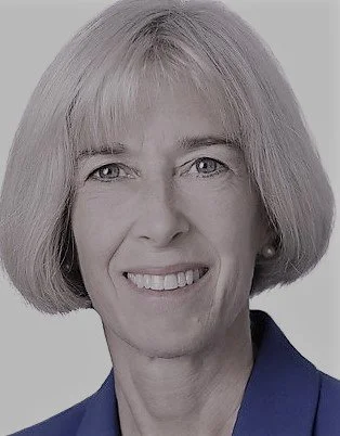 Close-up portrait of a woman with short blonde hair, smiling, wearing a dark blue blazer against a light background.