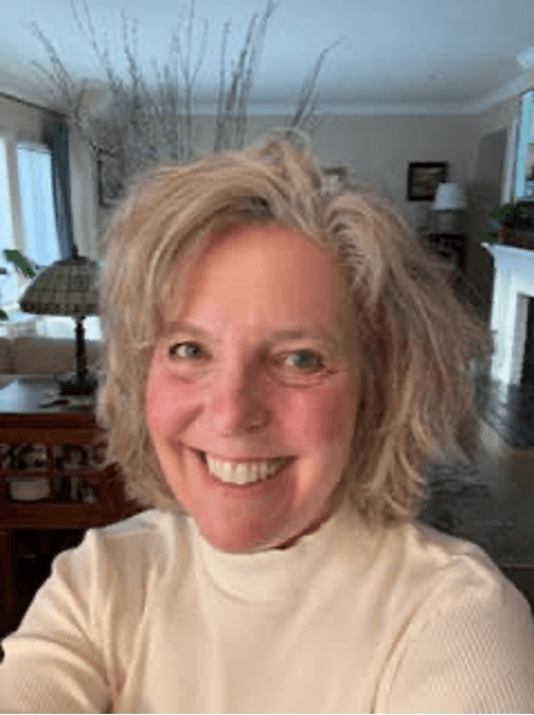 A woman with shoulder-length, curly blonde hair smiling in a living room with a sofa, lamp, and decorative items in the background.