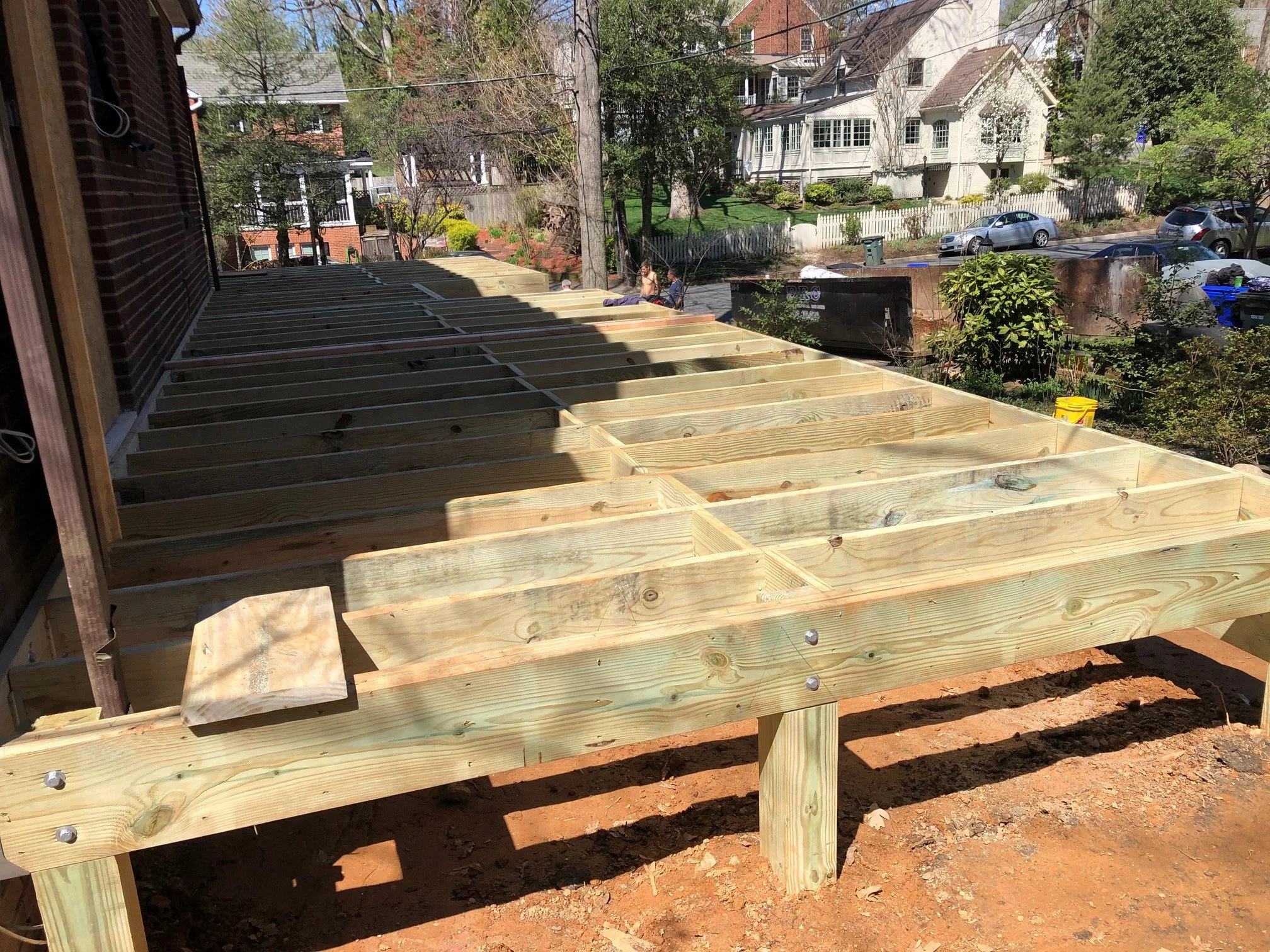 Wooden deck platform under construction on a residential backyard, with houses, trees, and parked cars in the background.