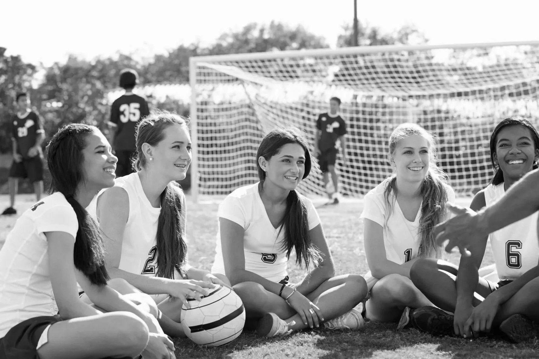 A group of female soccer players sitting on the grass and smiling, with a soccer ball in front of them and a soccer goal in the background.
