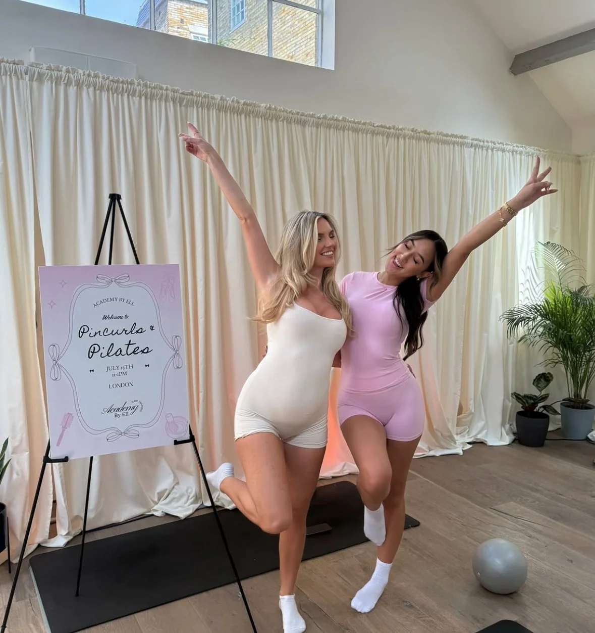Two women in workout attire dancing and smiling in a fitness studio with a sign promoting a Pintucks and Pilates class.
