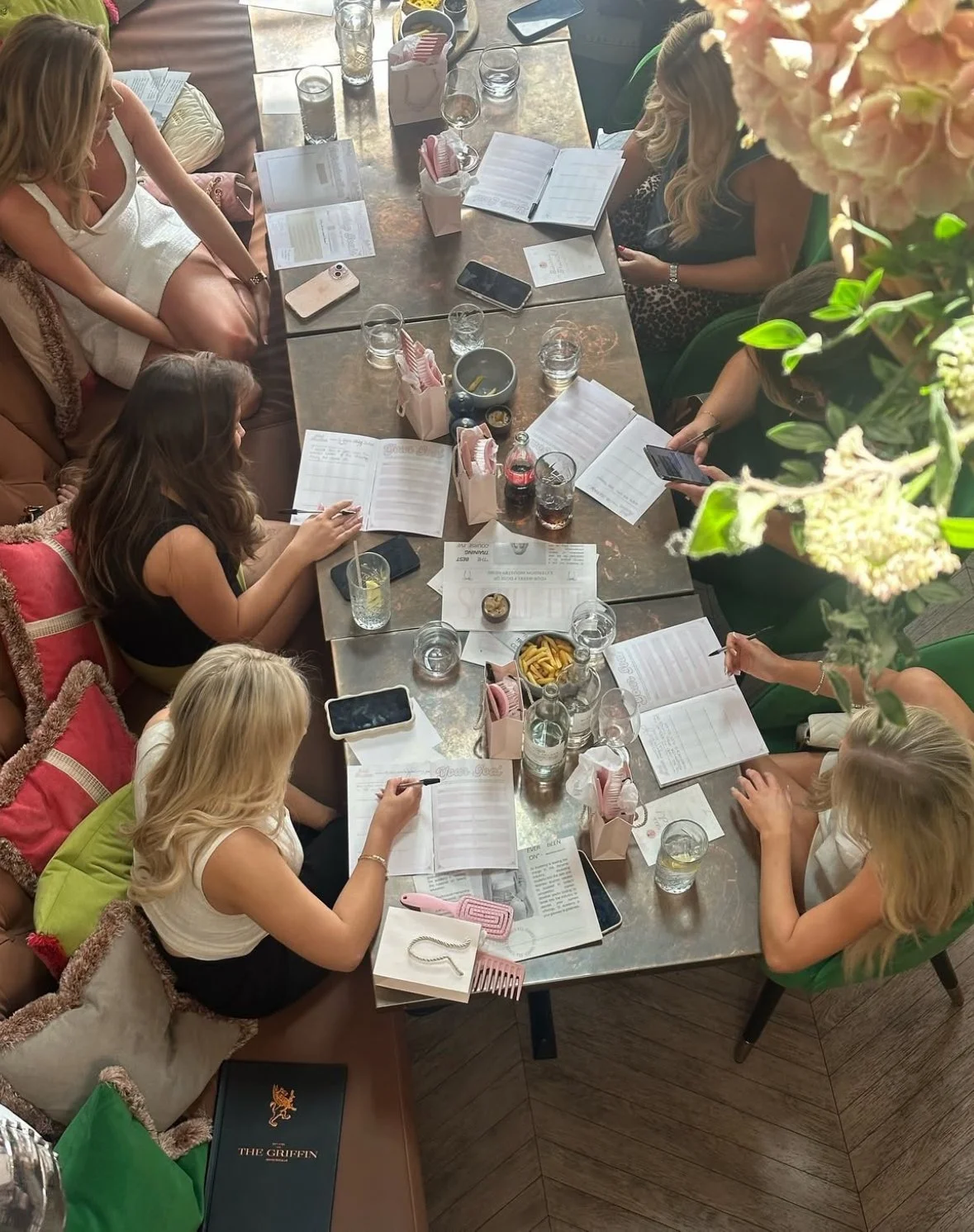 A top-down view of a group of women sitting around a long table in a restaurant or cafe, engaged in a meeting or gathering. The table is scattered with glasses of water, notebooks, menus, phones, and small pink gift bags. Some women are writing, whil