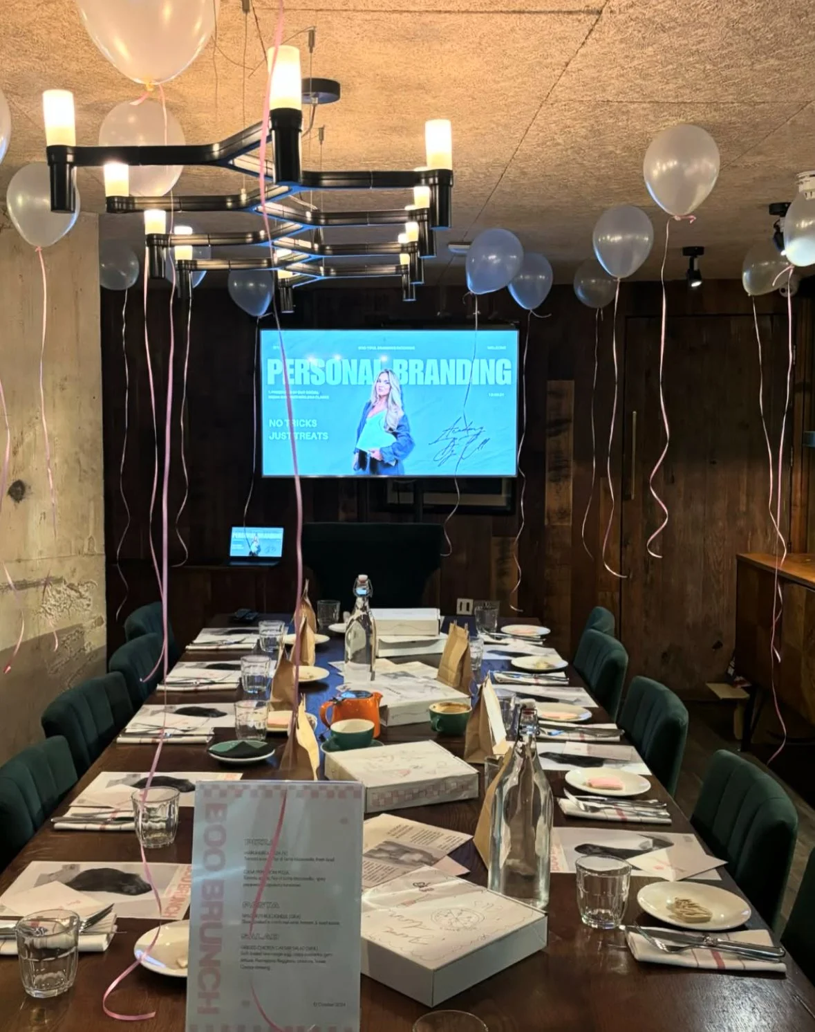 A long dining table set for a celebration with plates, glasses, napkins, and a boxed cake. White balloons with pink ribbons are tied to the chairs and hanging from the ceiling. A large screen on the wall displays a presentation titled 'Personal Brand