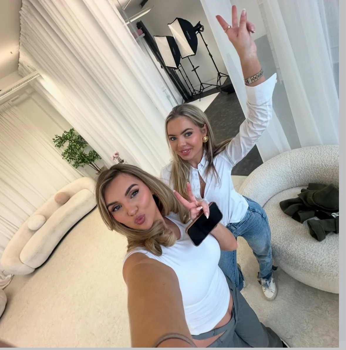 Two women taking a selfie in a studio or living room with photo equipment visible in the background. One woman is making a kissy face, and the other is smiling and holding a phone.
