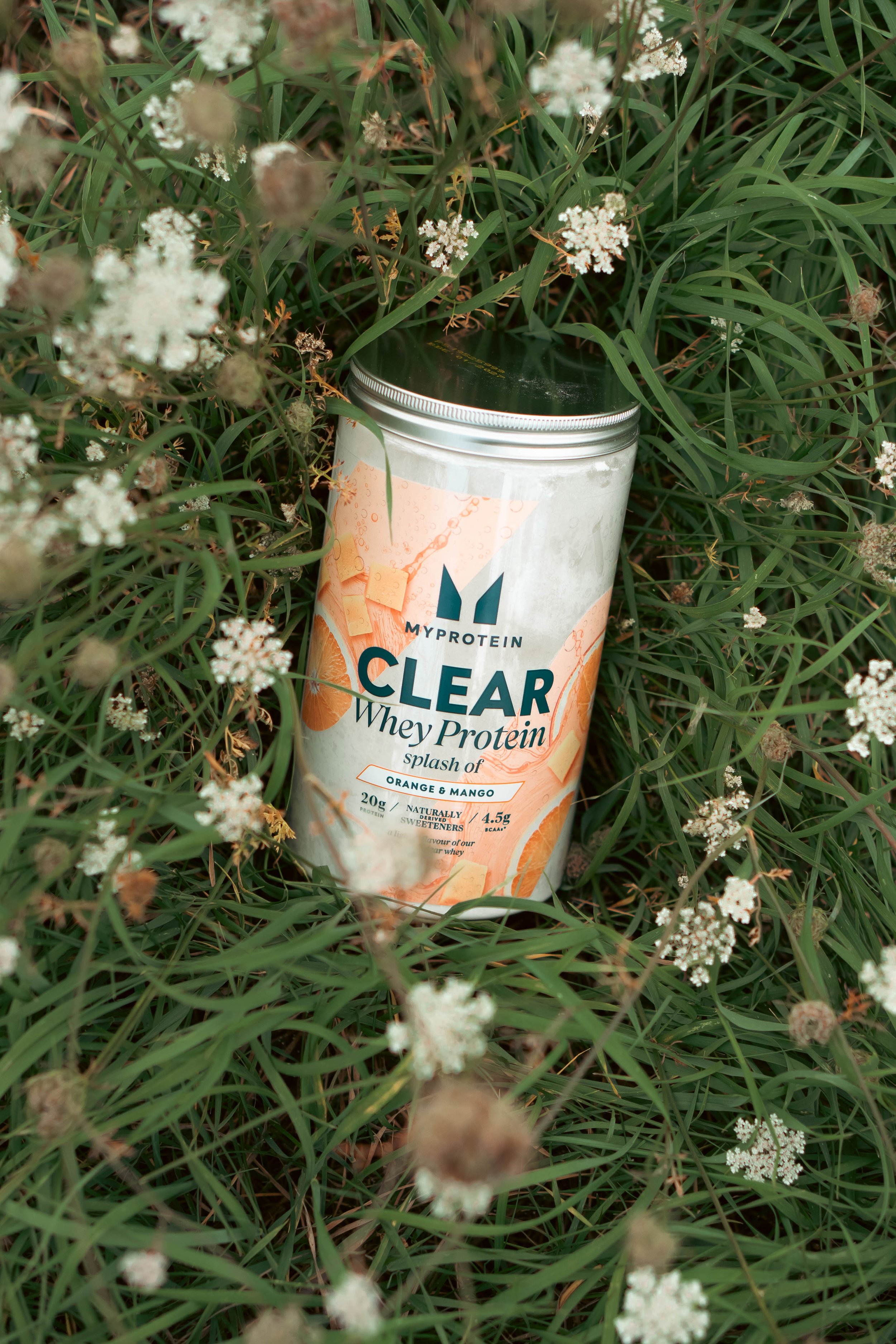 Een glas of blik met MYPROTEIN Clear Whey Protein splash of sinaasappel en mango, in het gras met kleine witte bloemen.