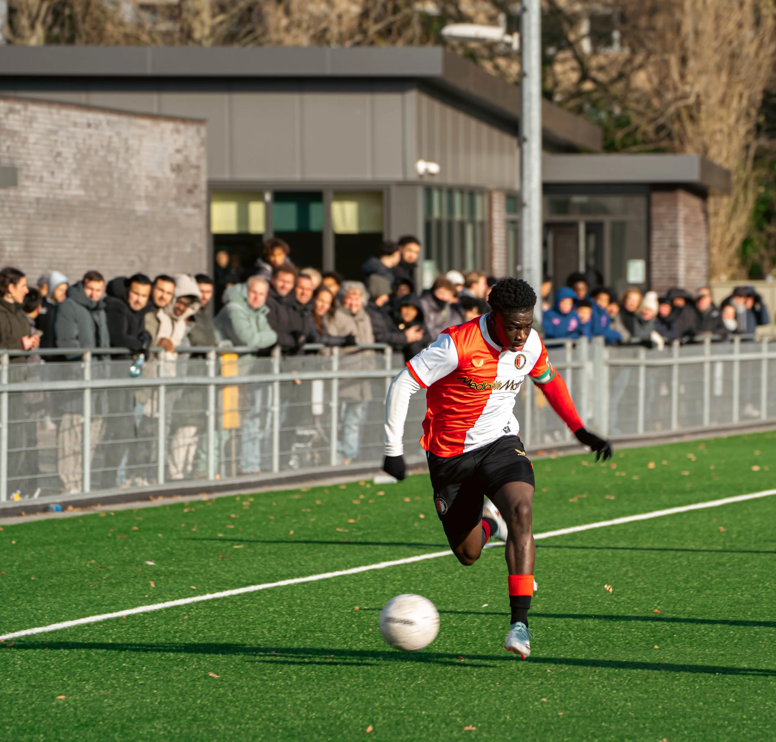 Voetballer in rood en wit shirt die de bal voortstuwt op een kunstgrasveld, met een rand van toeschouwers op de achtergrond.