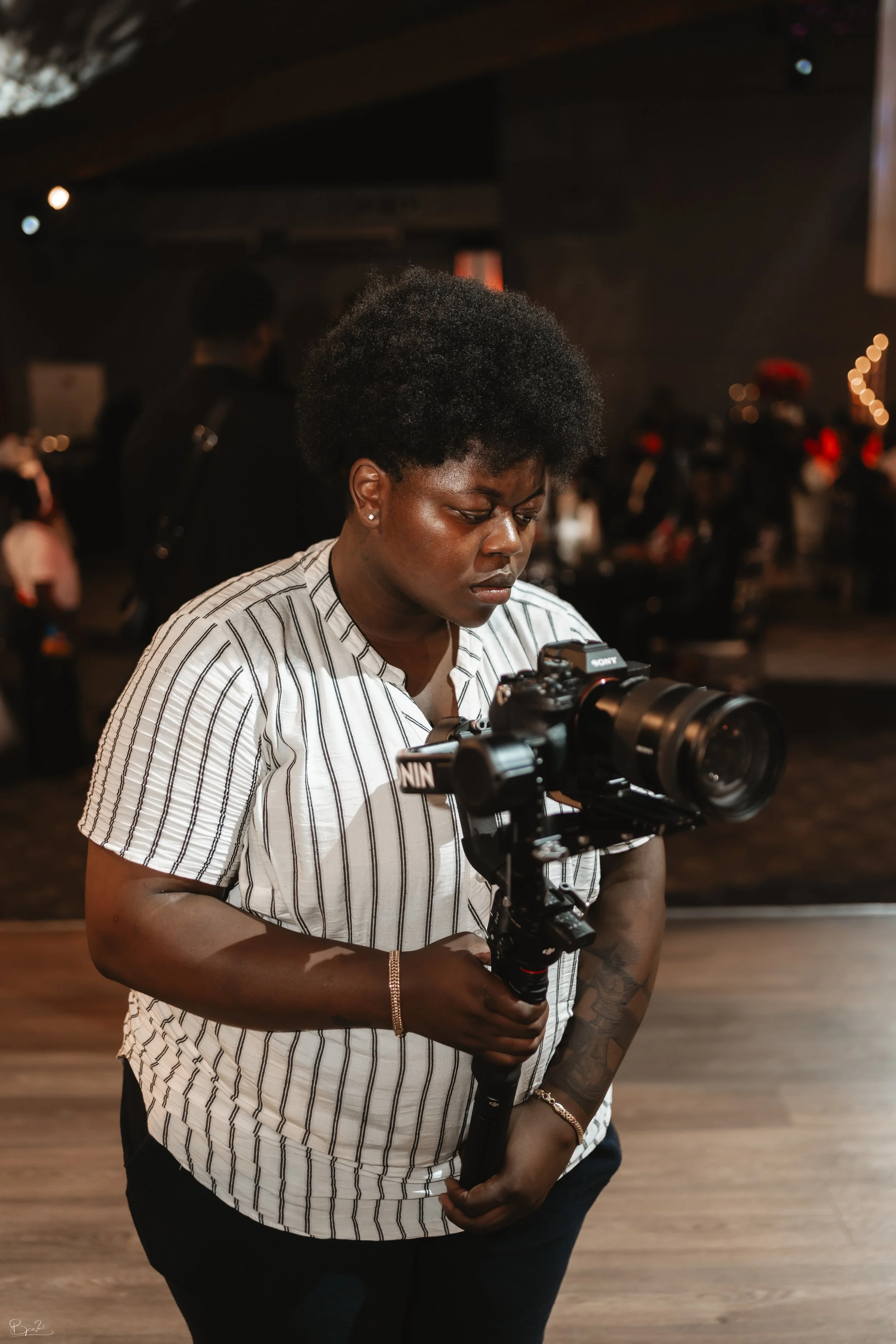 Een vrouw met een zwart krullend haar houdt een professionele camera met stabilisator vast in een indoor omgeving met meerdere mensen op de achtergrond.