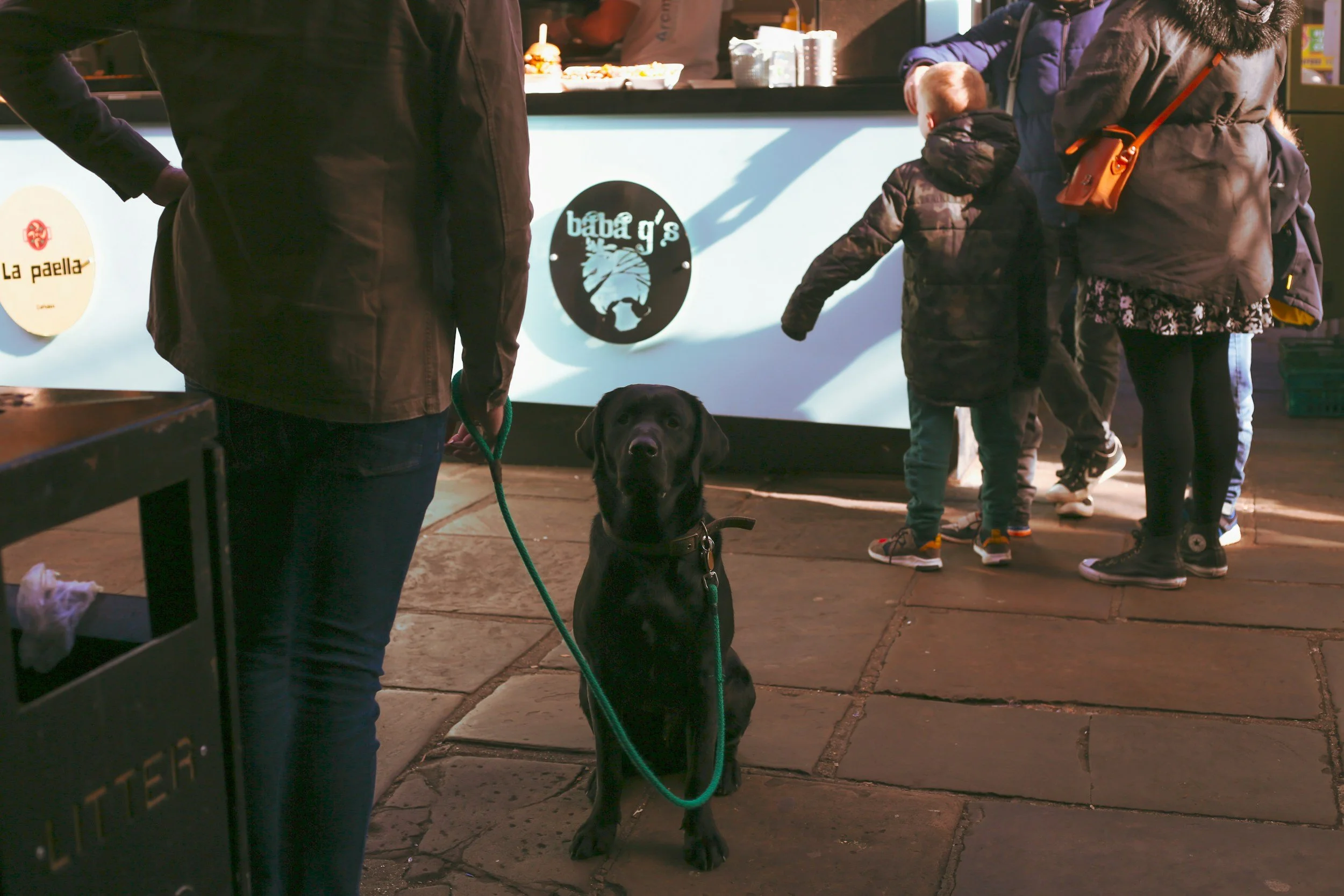 A black Labrador retriever sitting on a stone sidewalk in front of a food stand with people ordering food. The dog is on a green leash held by a person wearing dark jeans and a dark jacket. Several children and adults are near the stand, which has signs reading 'baba g's' and 'la paella'.