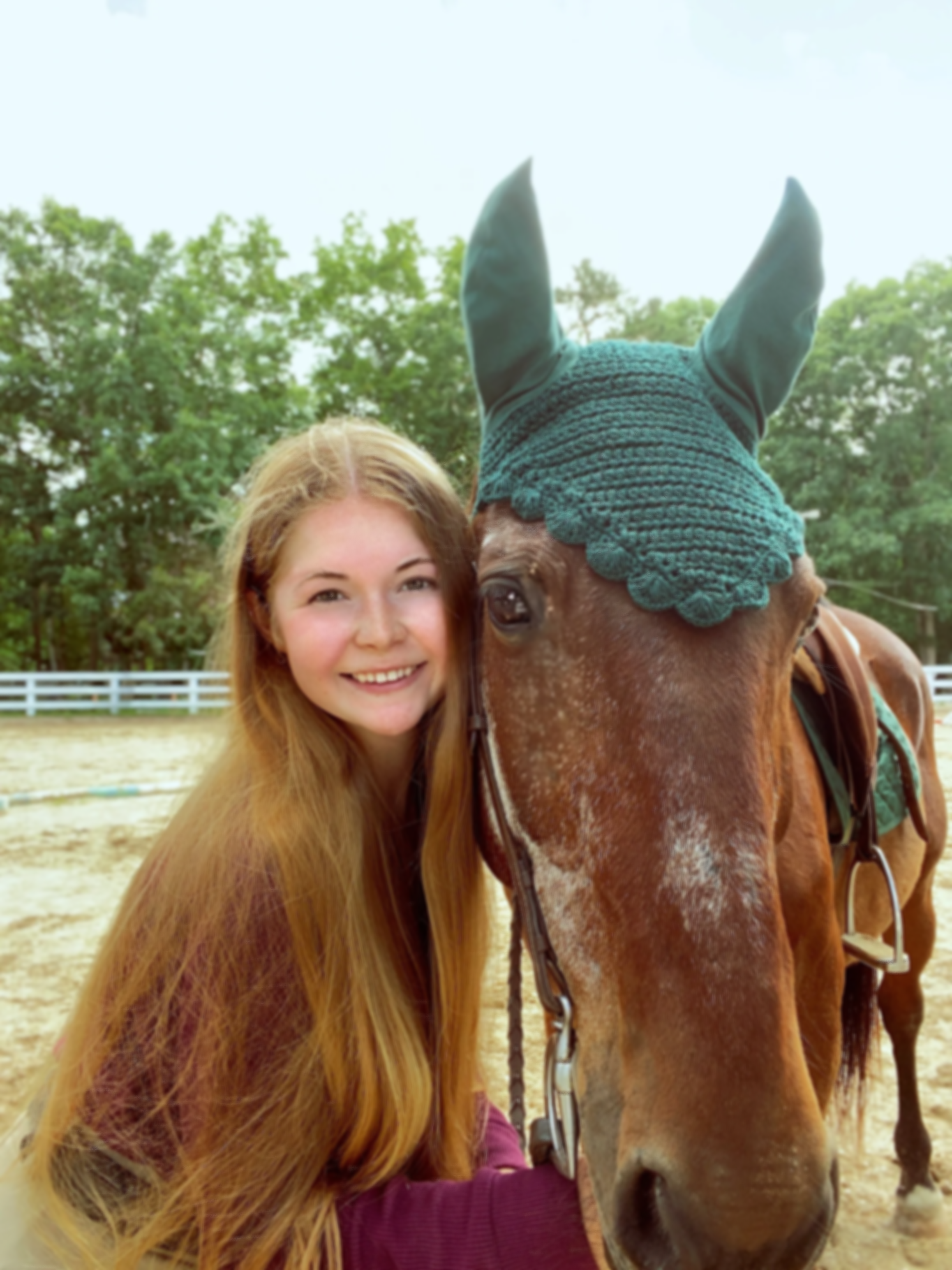 A young woman with long red hair smiling and standing close to a brown horse wearing a blue knit ear cover, outdoors in a paddock with trees and a white fence in the background.