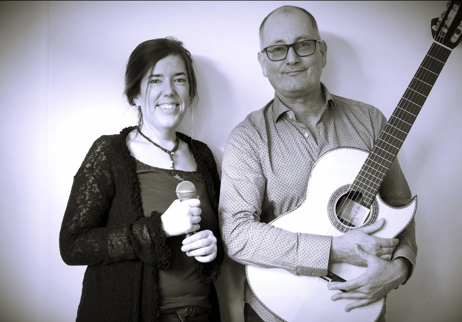 Ein Mann mit Brille hält eine Gitarre, eine Frau mit dunklem Haar und Kette hält ein Mikrofon, beide stehen vor weißer Wand.