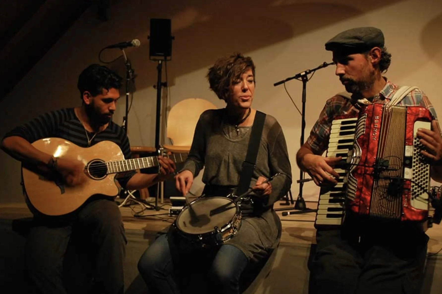 Drei Musiker spielen in einer kleinen Bühne, einer mit Gitarre, eine mit Tamburin und einer mit Akkordeon.