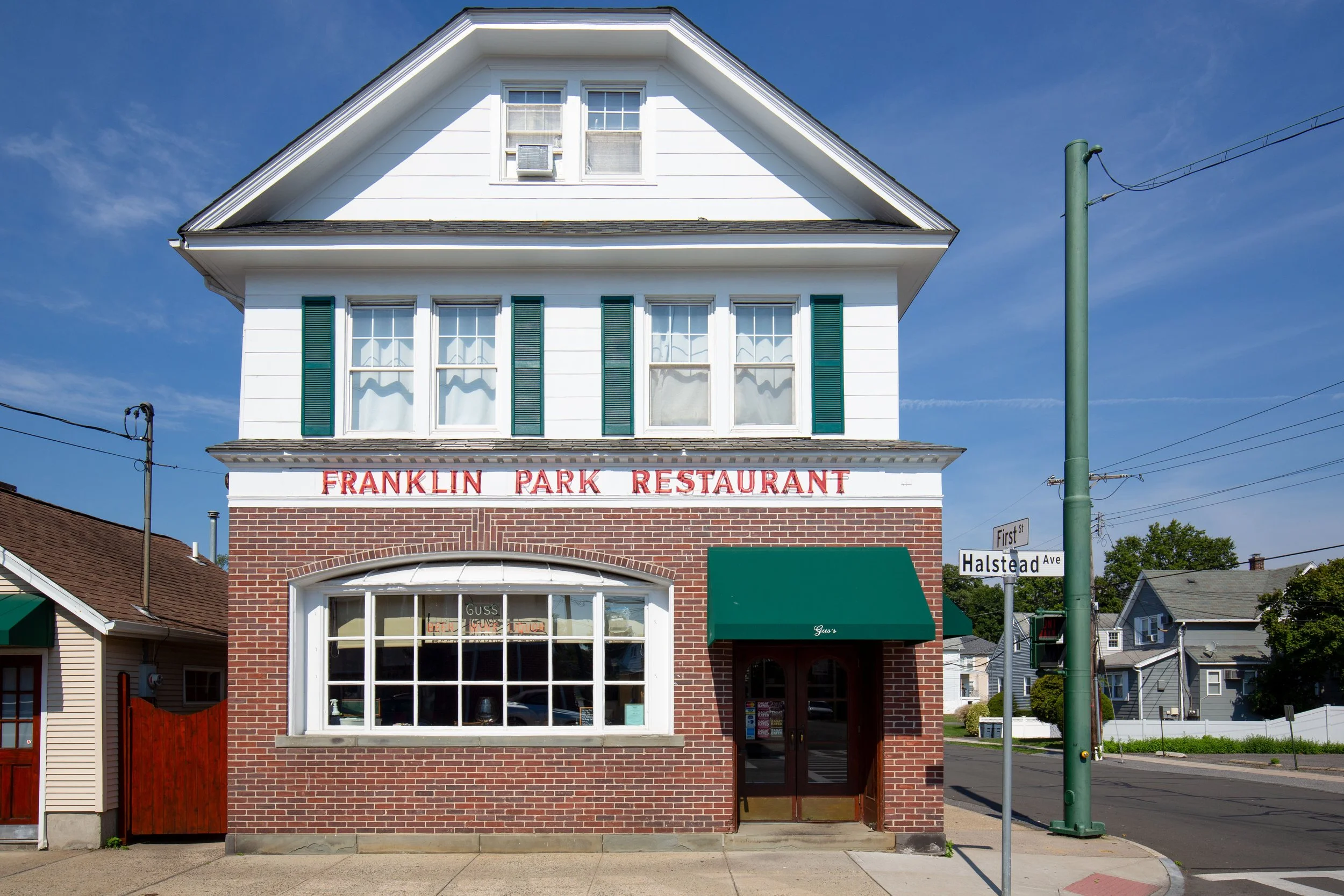 A three-story building with white siding and brick lower facade, housing the Franklin Park Restaurant, with a green awning over the entrance, located at a street corner on a sunny day.