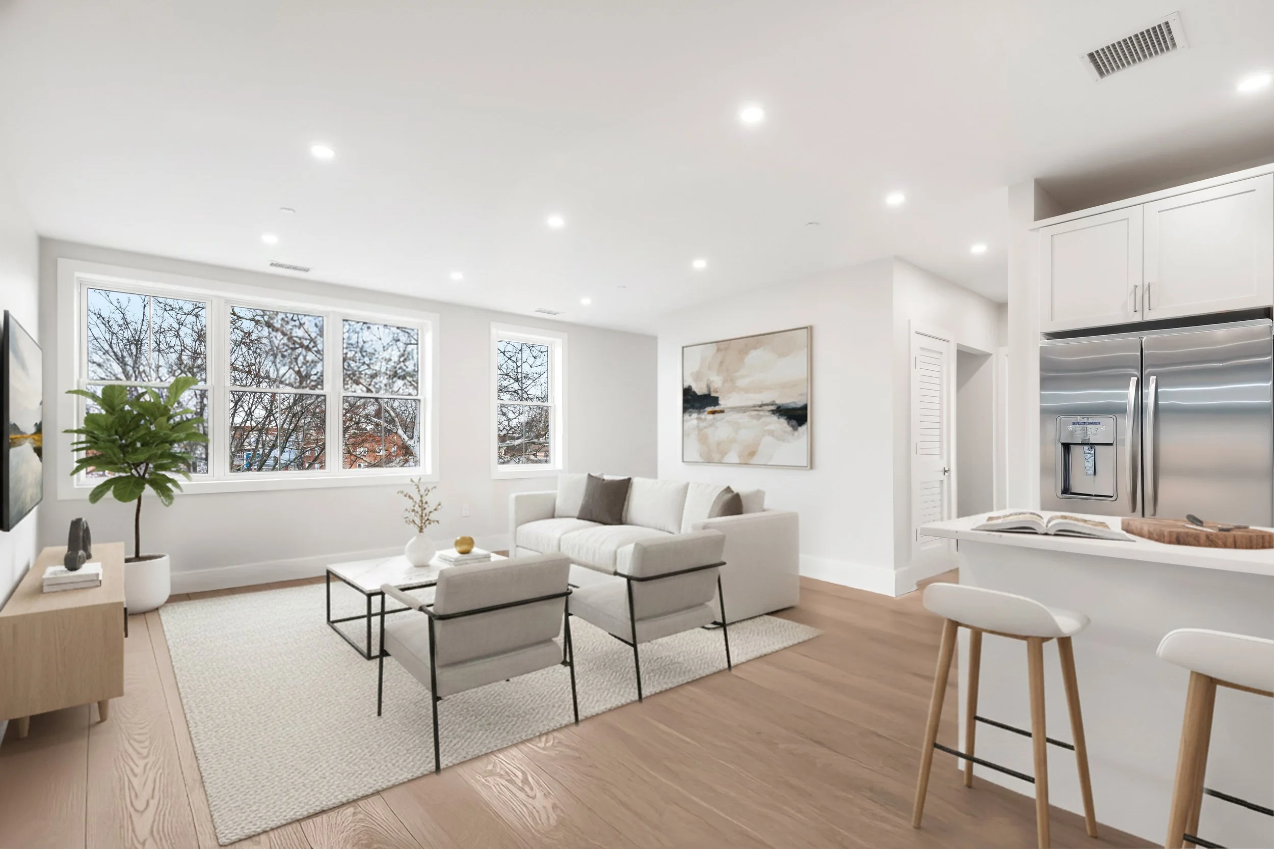 Bright living room with white walls and large windows, featuring a white sofa, artwork, potted plant, and a kitchen counter with stools.