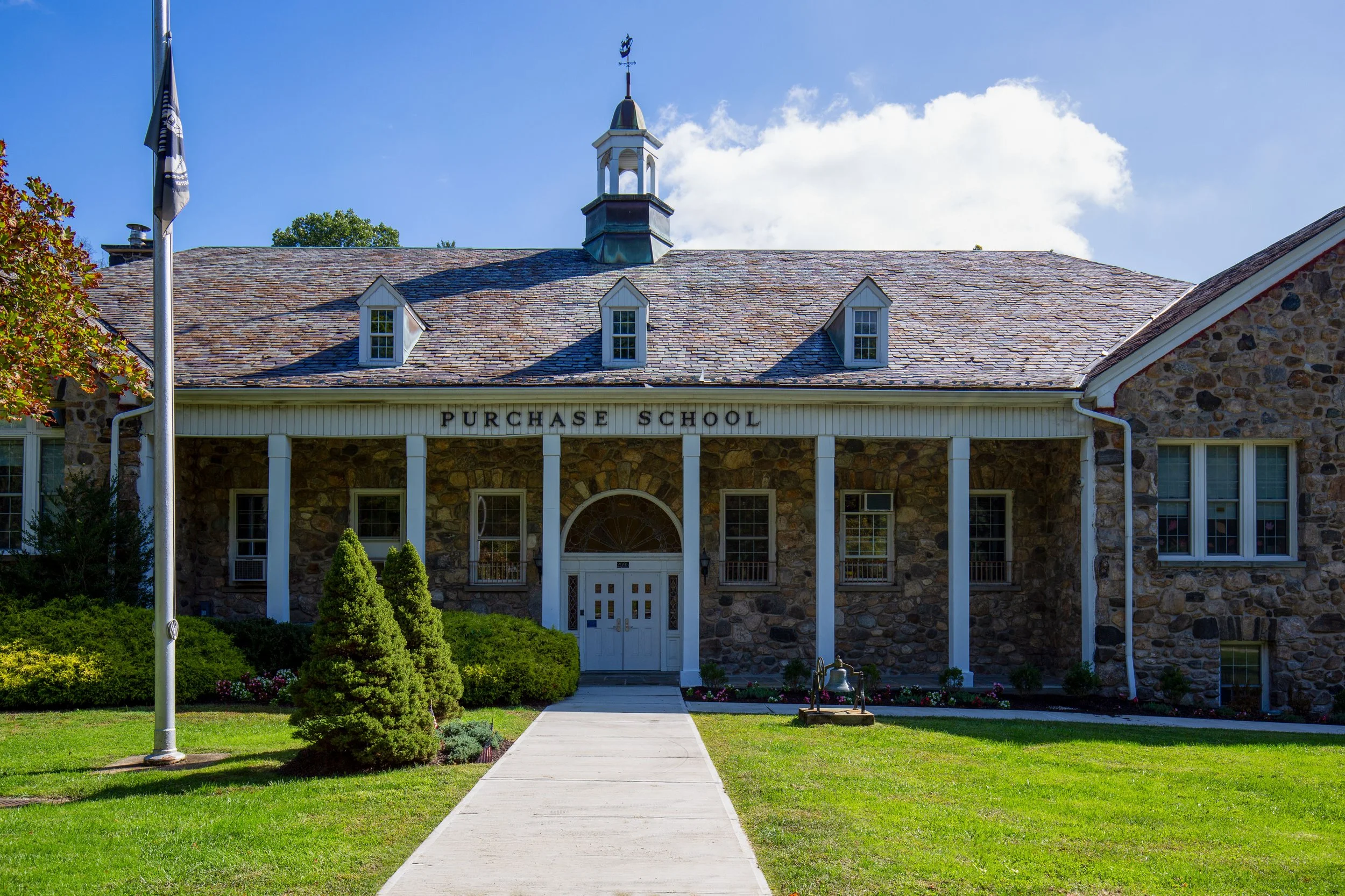 Front view of a stone school building with a sign that reads 'Purchase School,' a small courtyard, a flagpole, and a clear blue sky.