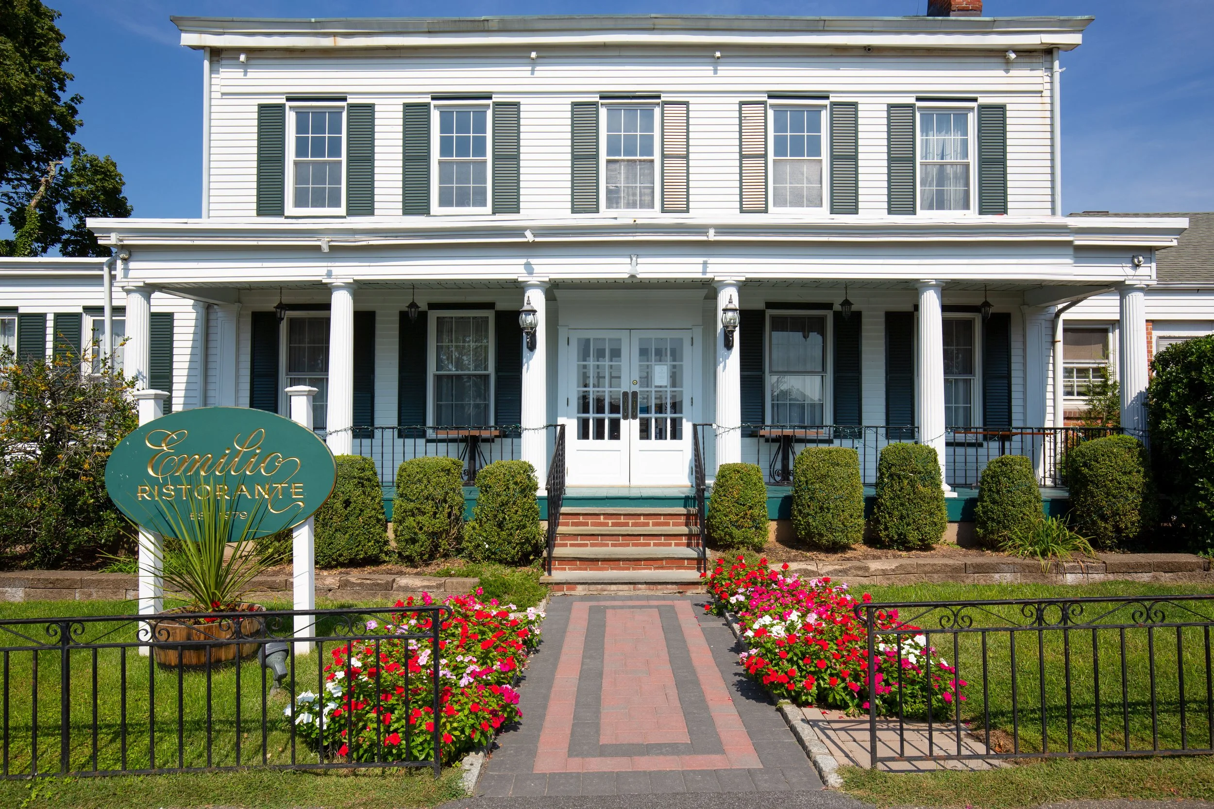 White two-story house with black shutters, front porch with columns, stairs leading up to white double doors, flower bed with pink and white flowers, green bushes, and a sign that says 'Emilio Ristorante' in front of the house.
