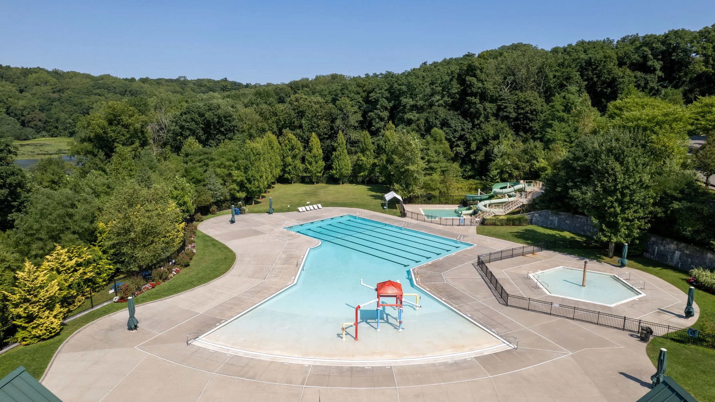 Empty outdoor swimming pool with shallow and deep sections, surrounded by a concrete deck, green trees, a grass lawn, and water slides in the background, under a clear blue sky.