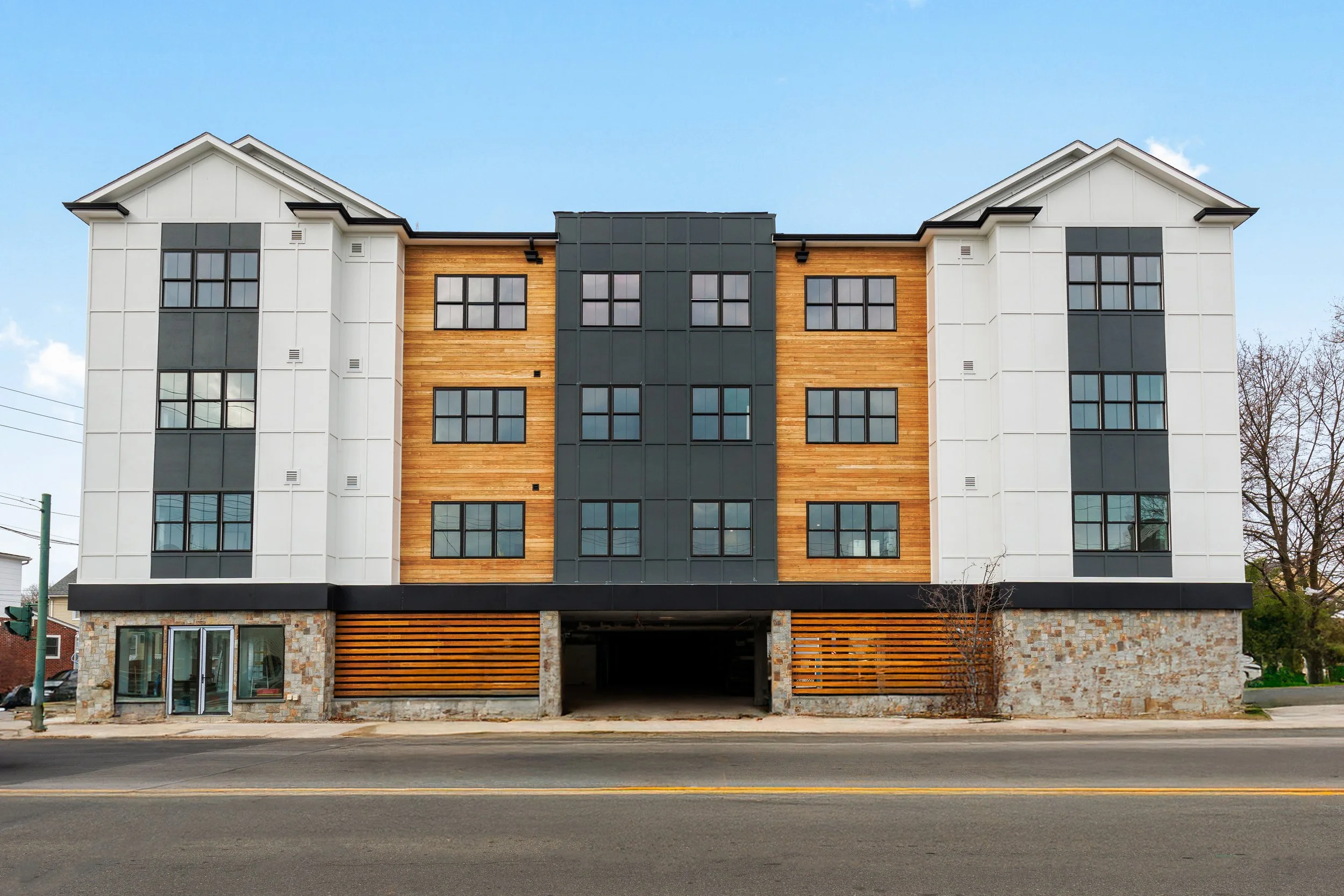 Front view of a modern multi-story residential building with white, black, and wooden exterior panels, large windows, and stonework at the ground level.