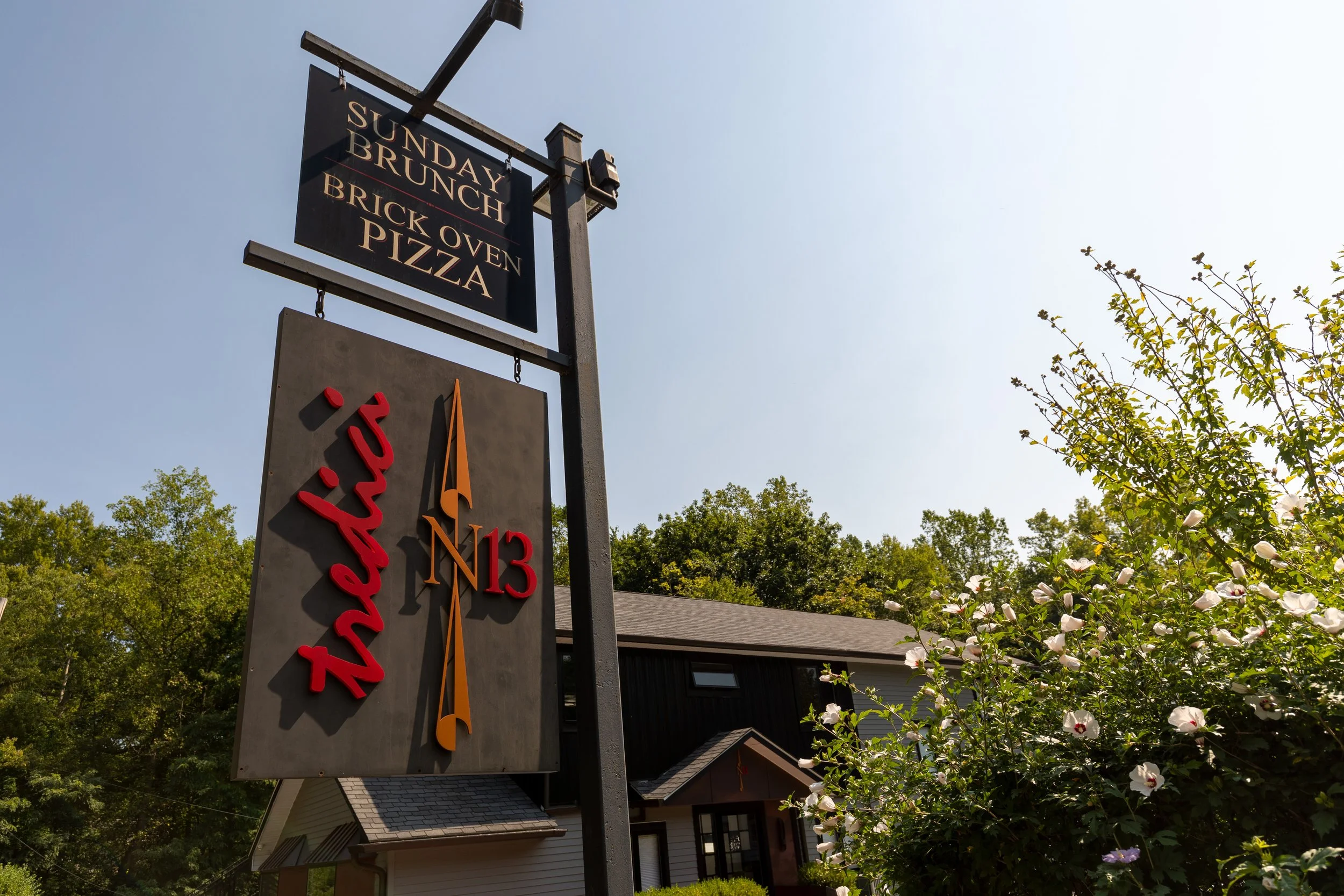 Signage for Thistle restaurant with a clock, showing 11:15, and a sign above advertising Sunday brunch, brick oven pizza, and a location on North 13th Street, with a house, trees, and flowering bushes in the background under a clear blue sky.