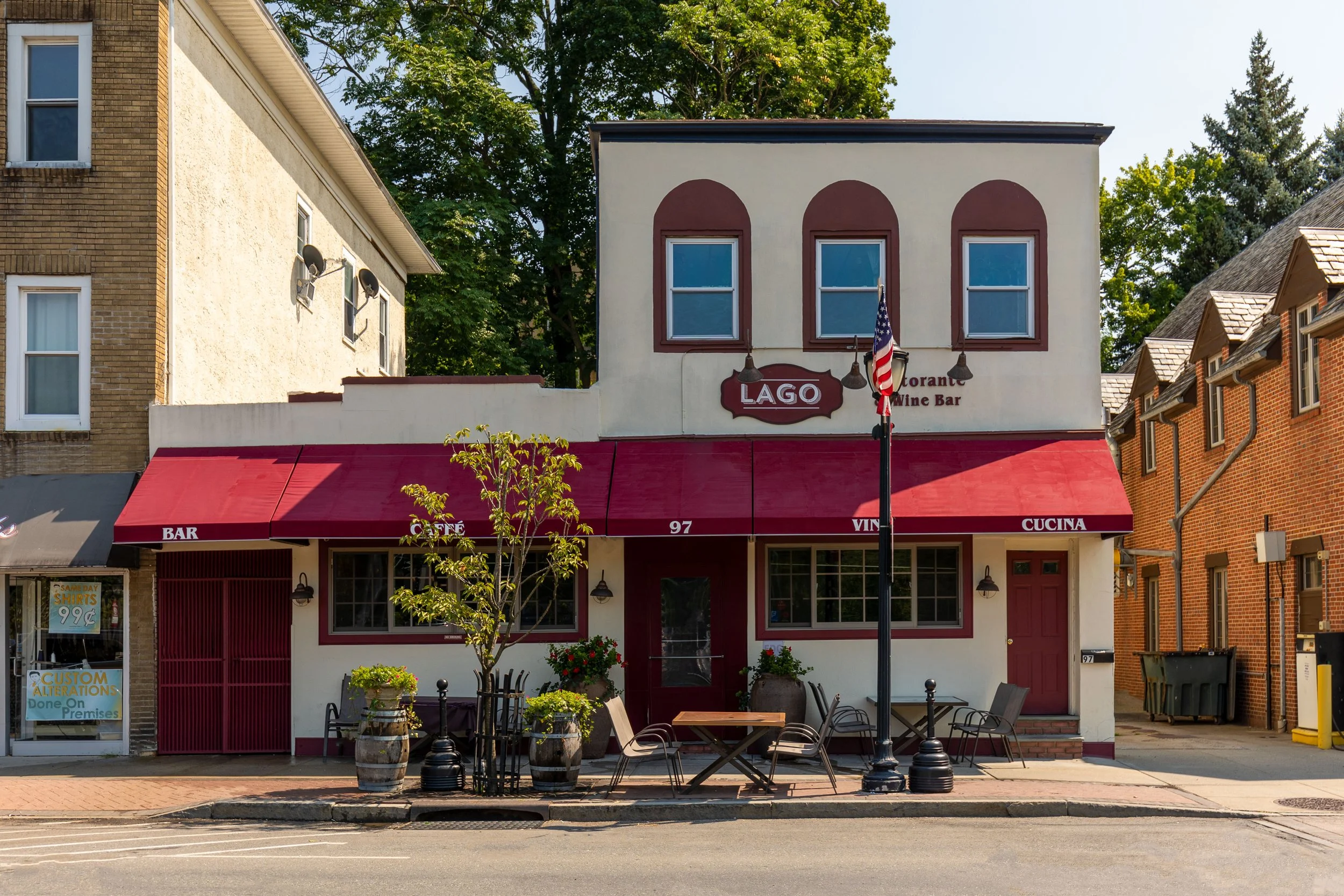 A two-story building with a white facade and red accents, featuring a restaurant named LAGO, with outdoor seating on the sidewalk, potted plants, a flagpole with an American flag, and other brick buildings on either side.