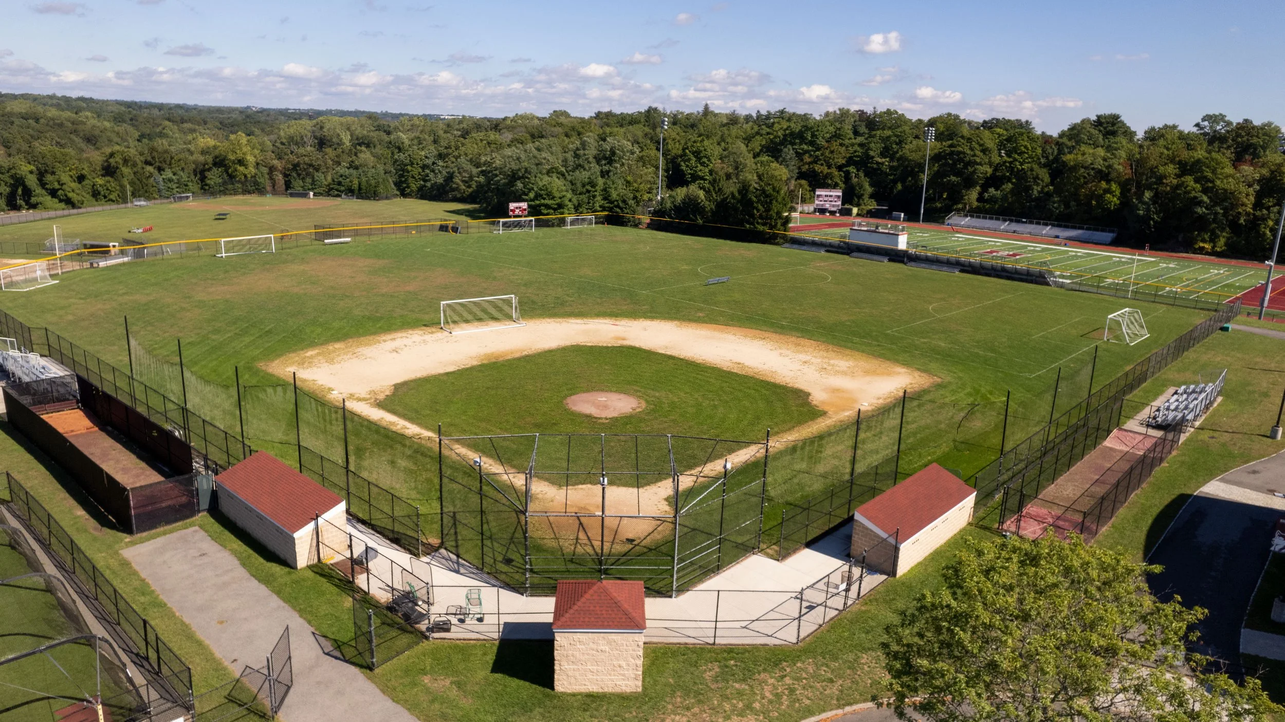 Aerial view of a sports complex featuring a baseball field in the foreground, multiple soccer fields in the middle ground, and a track and field stadium in the background, surrounded by trees and clear skies.