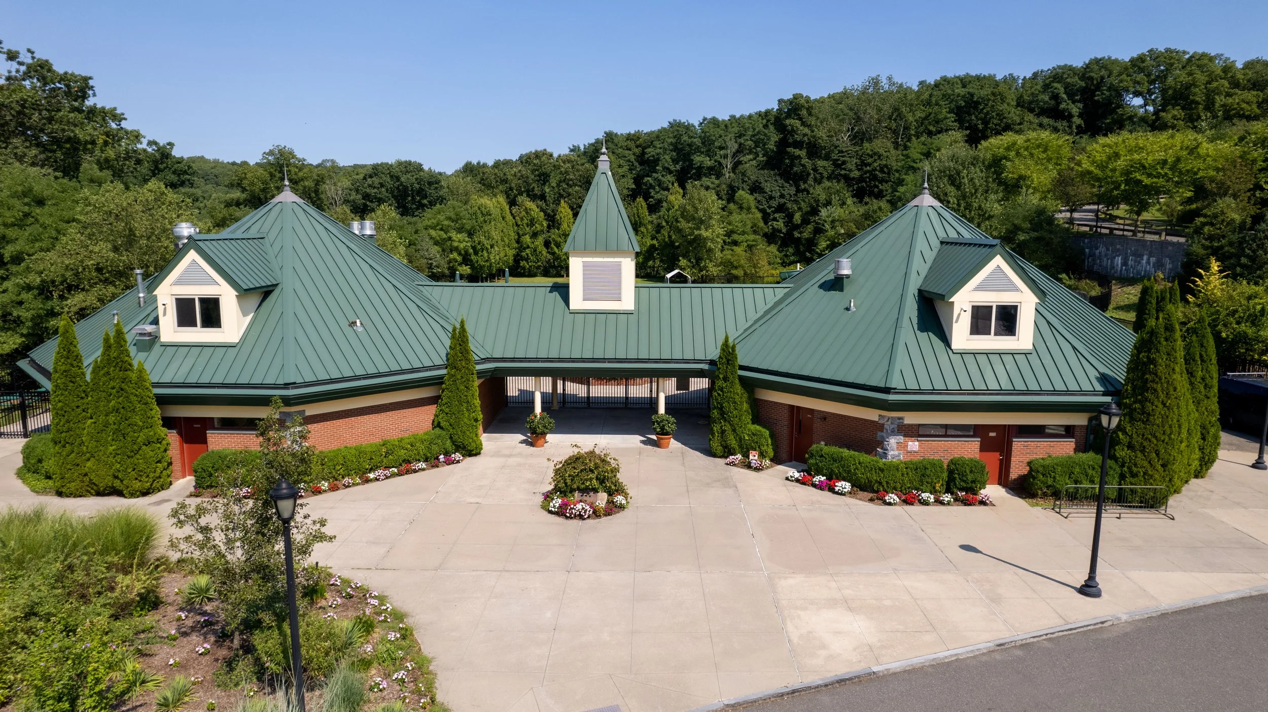 The image shows a large building with a green metal roof and brick walls, surrounded by landscaped bushes and flowers. There are two lamp posts in the front and a paved area leading to the entrance, with a backdrop of a tree-covered hill under a clea