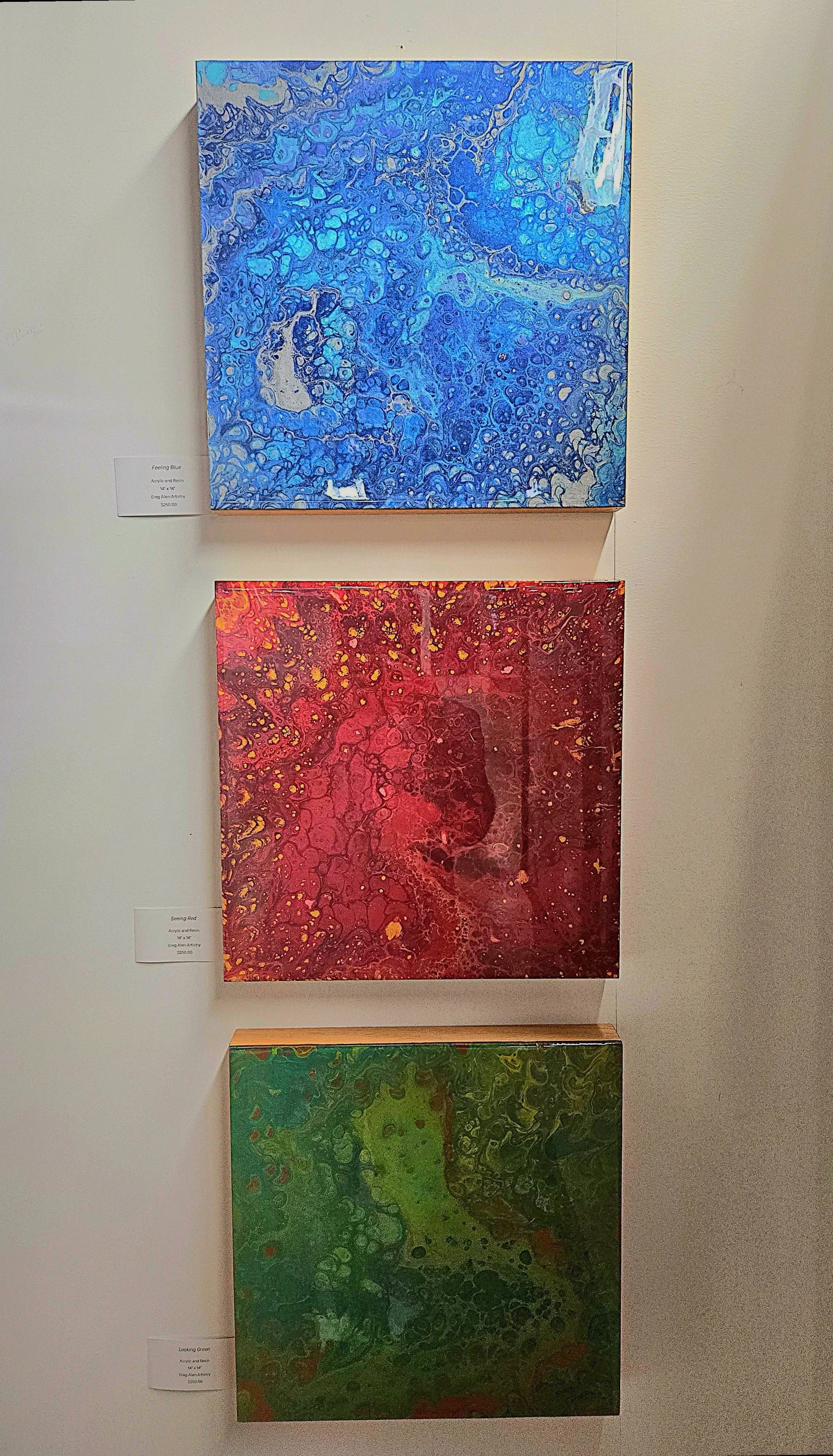 Three colorful abstract paintings on a gallery wall. The top painting features shades of blue with a bubble-like pattern. The middle painting displays red and yellow with a fluid, organic design. The bottom painting shows green hues with a similar fluid pattern.