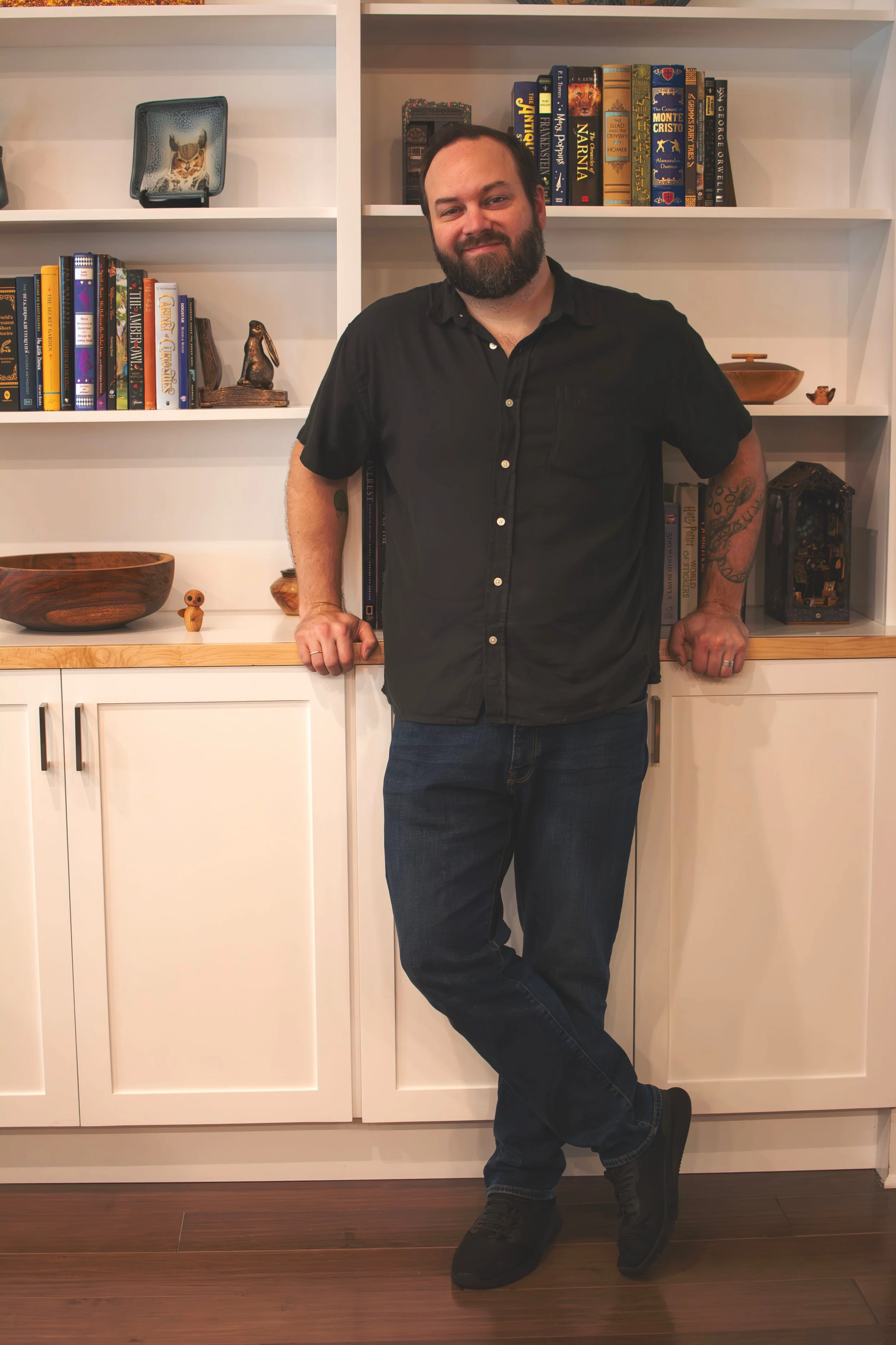 A man with a beard and tattoos on his left arm standing in front of a white bookshelf filled with books and decorative items. He is wearing a black short-sleeved button-up shirt and dark jeans, with his right hand resting on the lower cabinet and his left hand on the shelf. He has a relaxed stance with one leg crossed over the other at the ankle.
