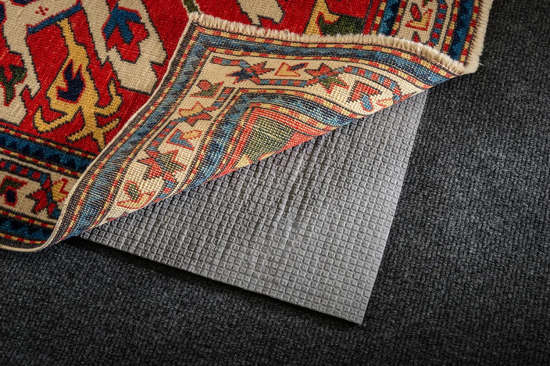 Close-up of a corner of a patterned area rug lifted to reveal a gray, textured rubber mat underneath, on a dark carpeted floor.