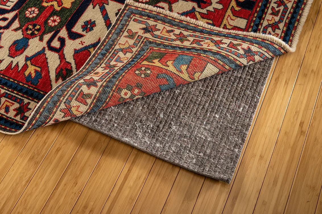 A patterned area rug partially rolled back to reveal a gray underlay, on a wooden floor.