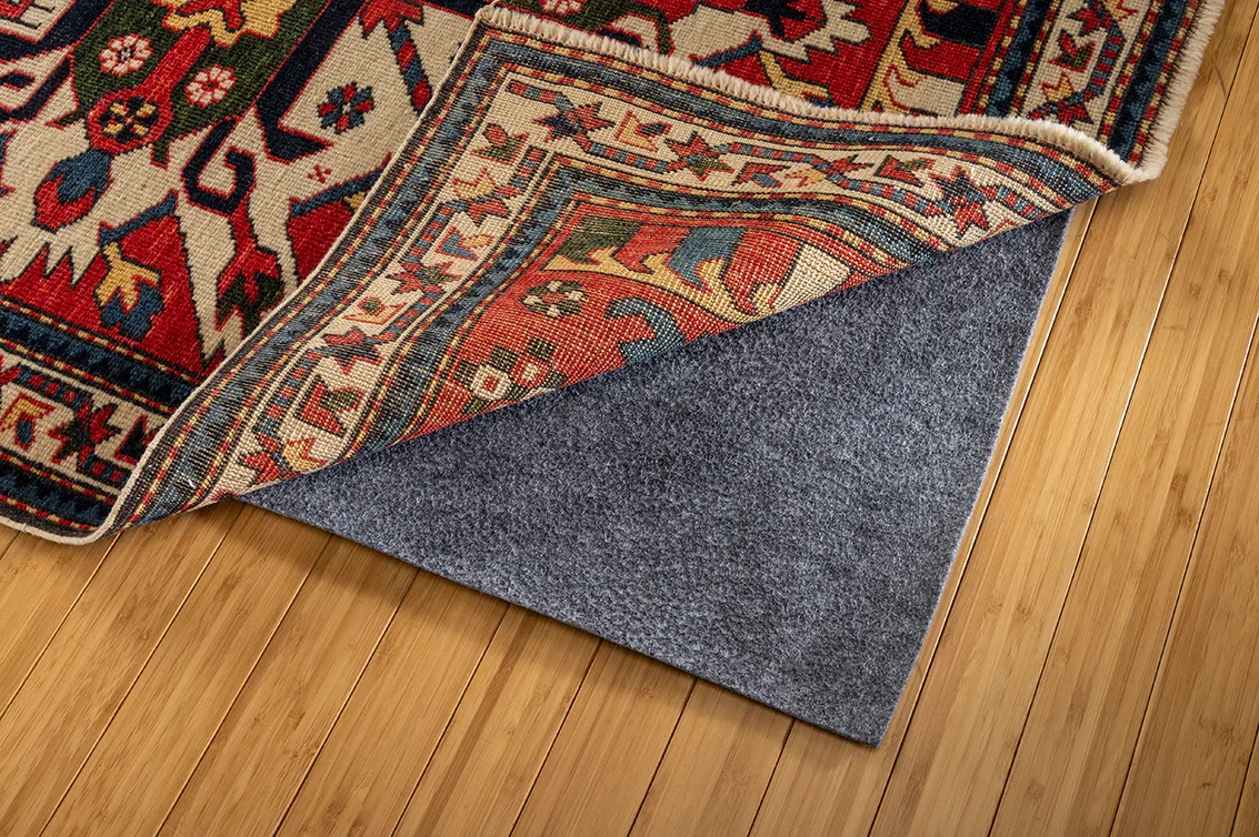 Close-up of a patterned area rug partially lifted to reveal a gray non-slip rug pad underneath, on a hardwood floor.
