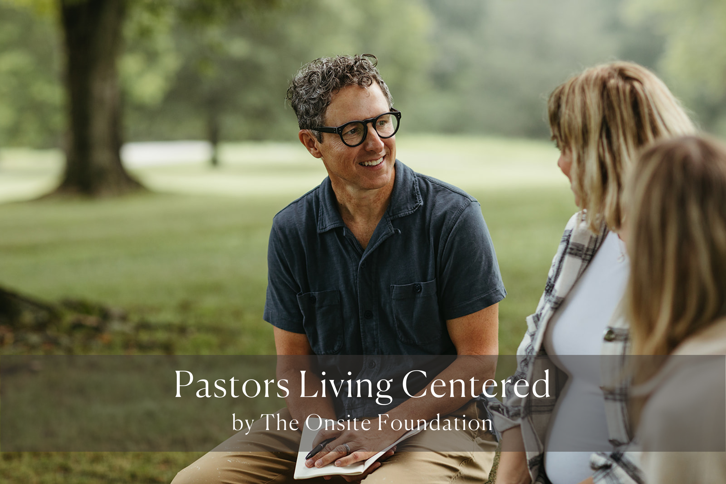 A man with curly hair and glasses talking to two women outdoors in a park setting.