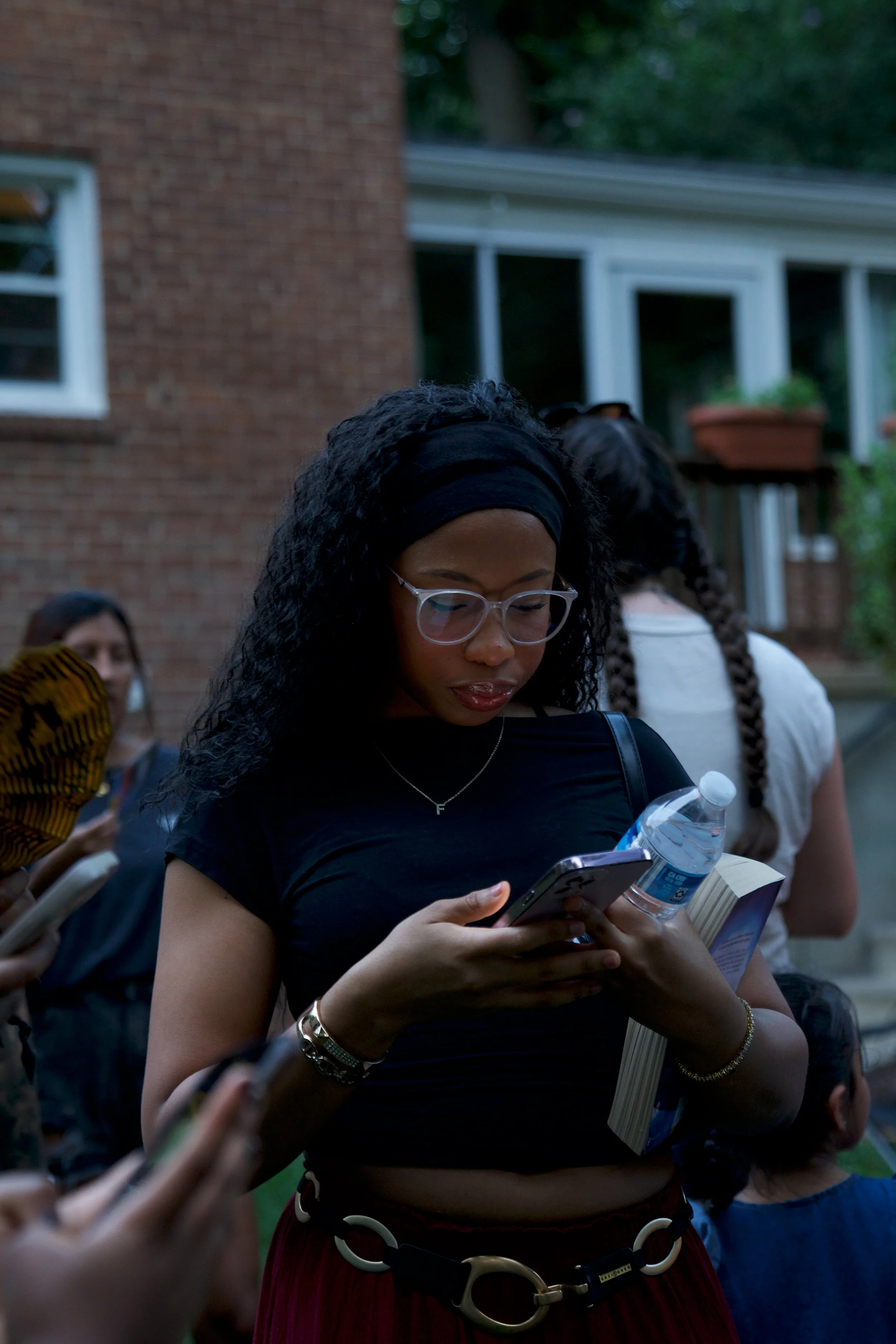A young woman with curly black hair, glasses, and a black headband looking at her phone while holding a water bottle and a book outdoors, with other people in the background.