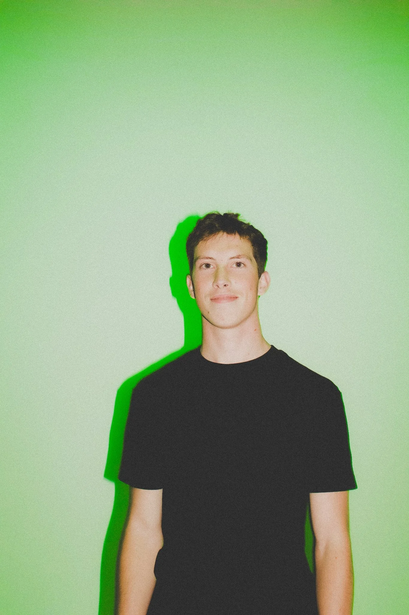 Young man with short dark hair wearing a black t-shirt, standing against a light green wall, with a green shadow cast on the wall behind him.