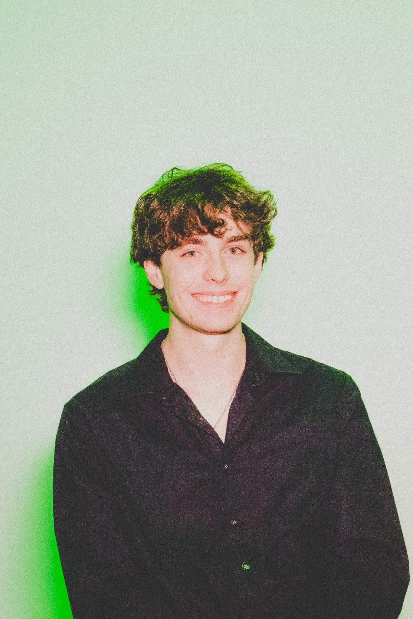 A young man with curly brown hair, light skin, and a bright smile, wearing a black shirt, standing against a white background with green lighting on one side.