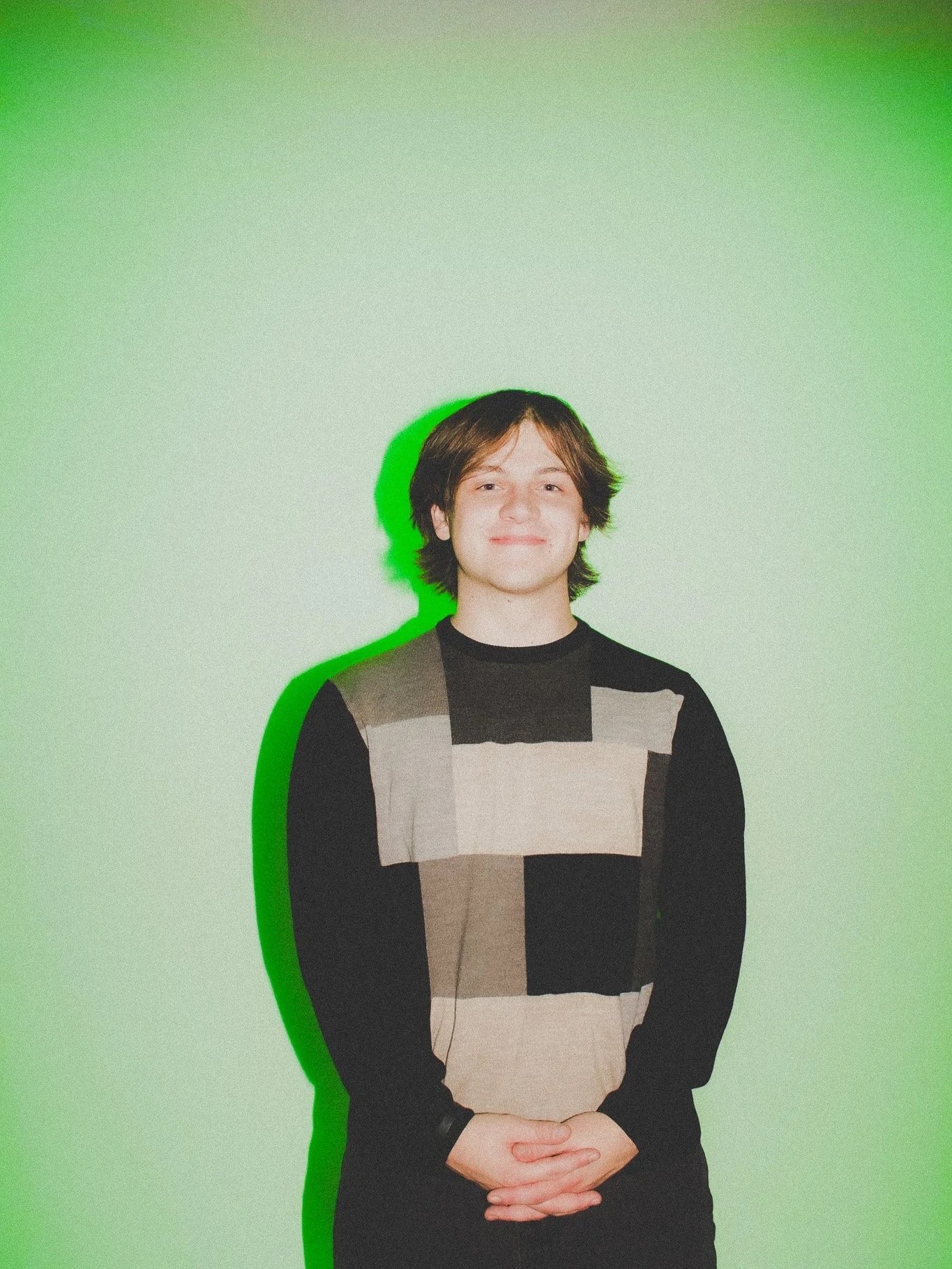 Person with medium-length brown hair and light skin standing against a green background, wearing a black and beige color-block sweater, with a green shadow effect.