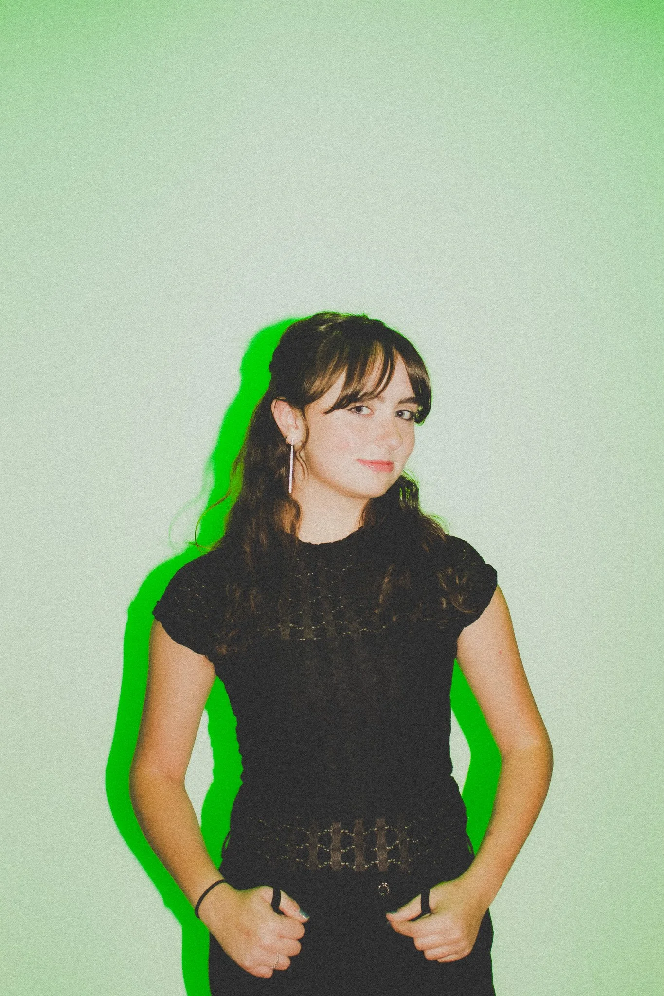 A young woman with long wavy hair wearing a black lace top, standing against a green background with a green shadow effect.
