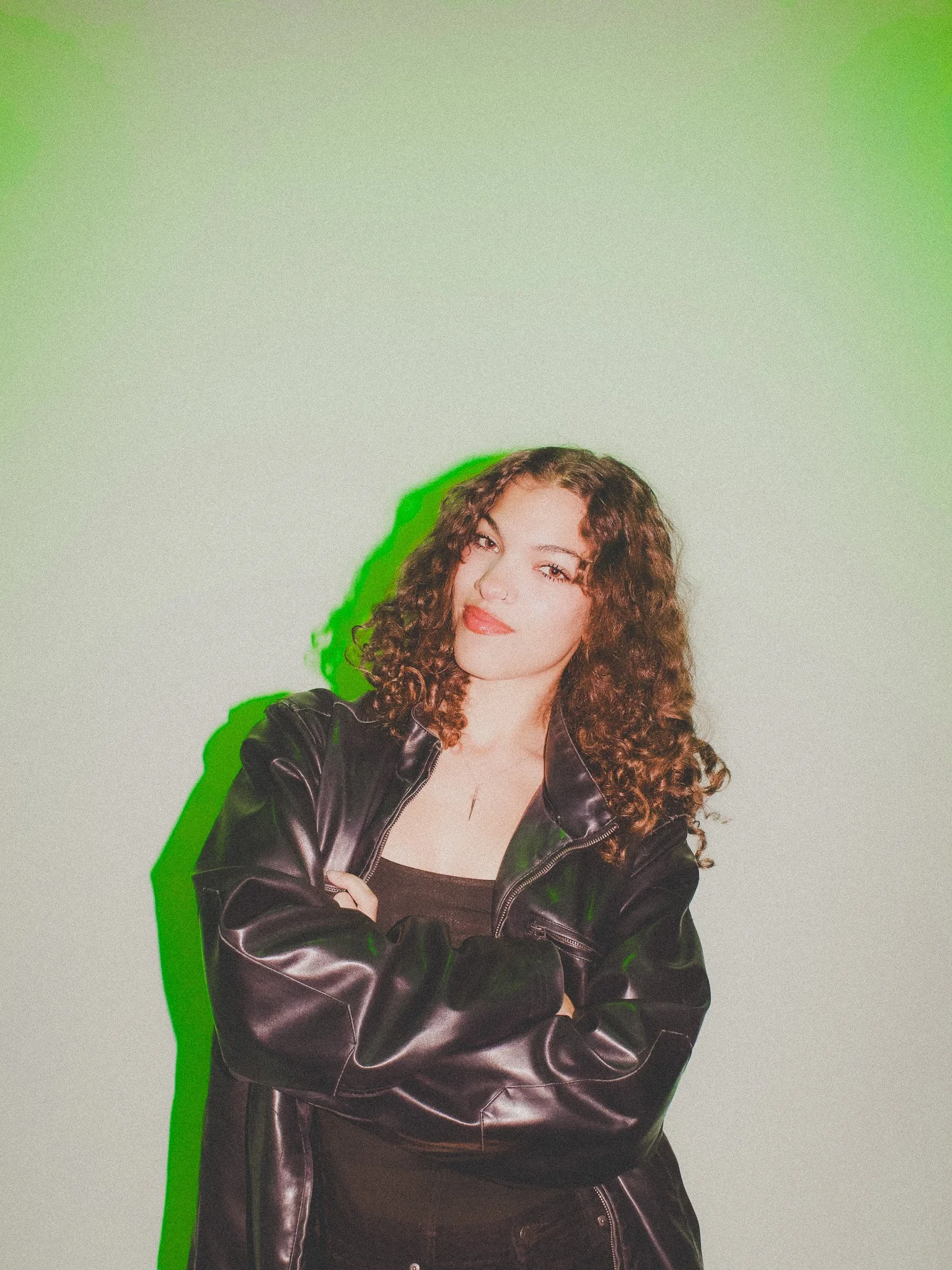 A young woman with curly hair wearing a black leather jacket and black top, standing with arms crossed against a plain background with a green shadow effect.
