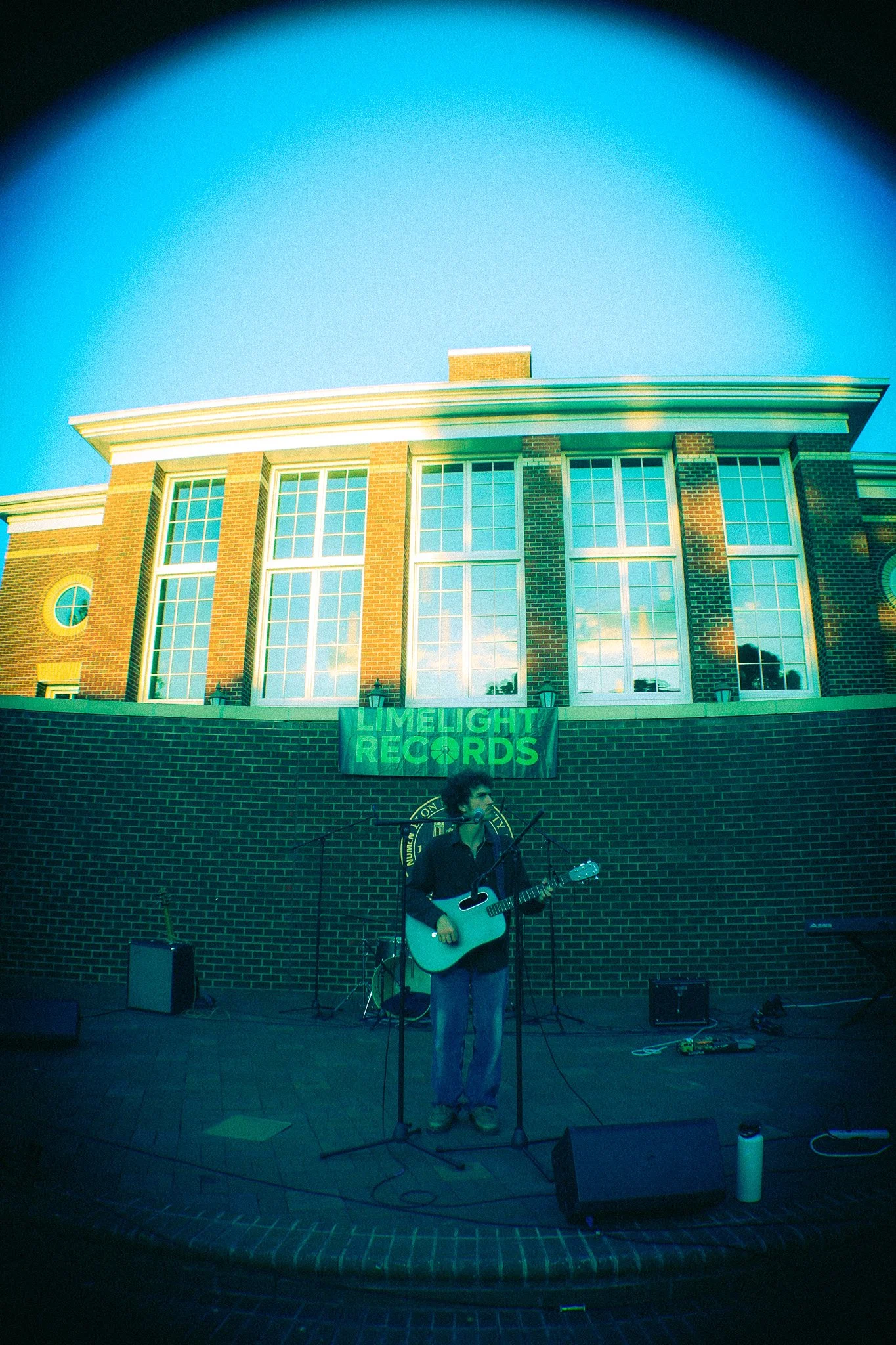 A musician with curly hair playing an acoustic guitar on an outdoor stage at night, with a brick building and a green 'LIMEIGHT RECORDS' banner hanging on the wall behind him.
