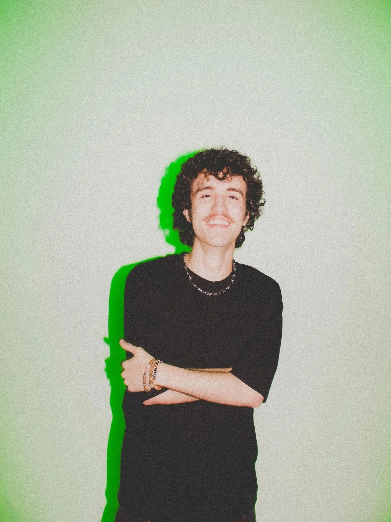 Young man with curly hair, mustache, wearing a black shirt and jewelry, smiling with arms crossed, standing against a light background with a green shadow effect.