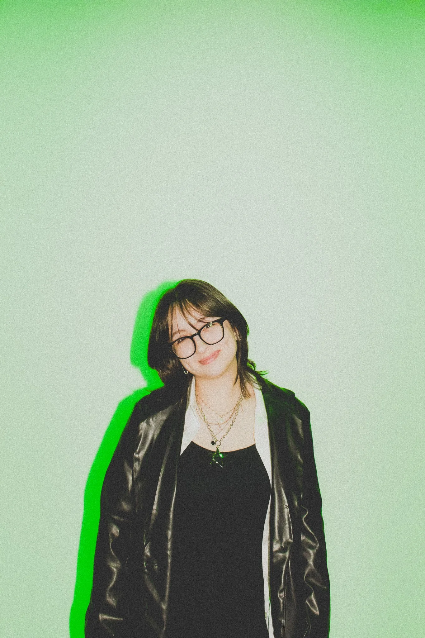 A young woman with black hair, glasses, and layered necklaces smiling against a light green background with a green shadow to her right.