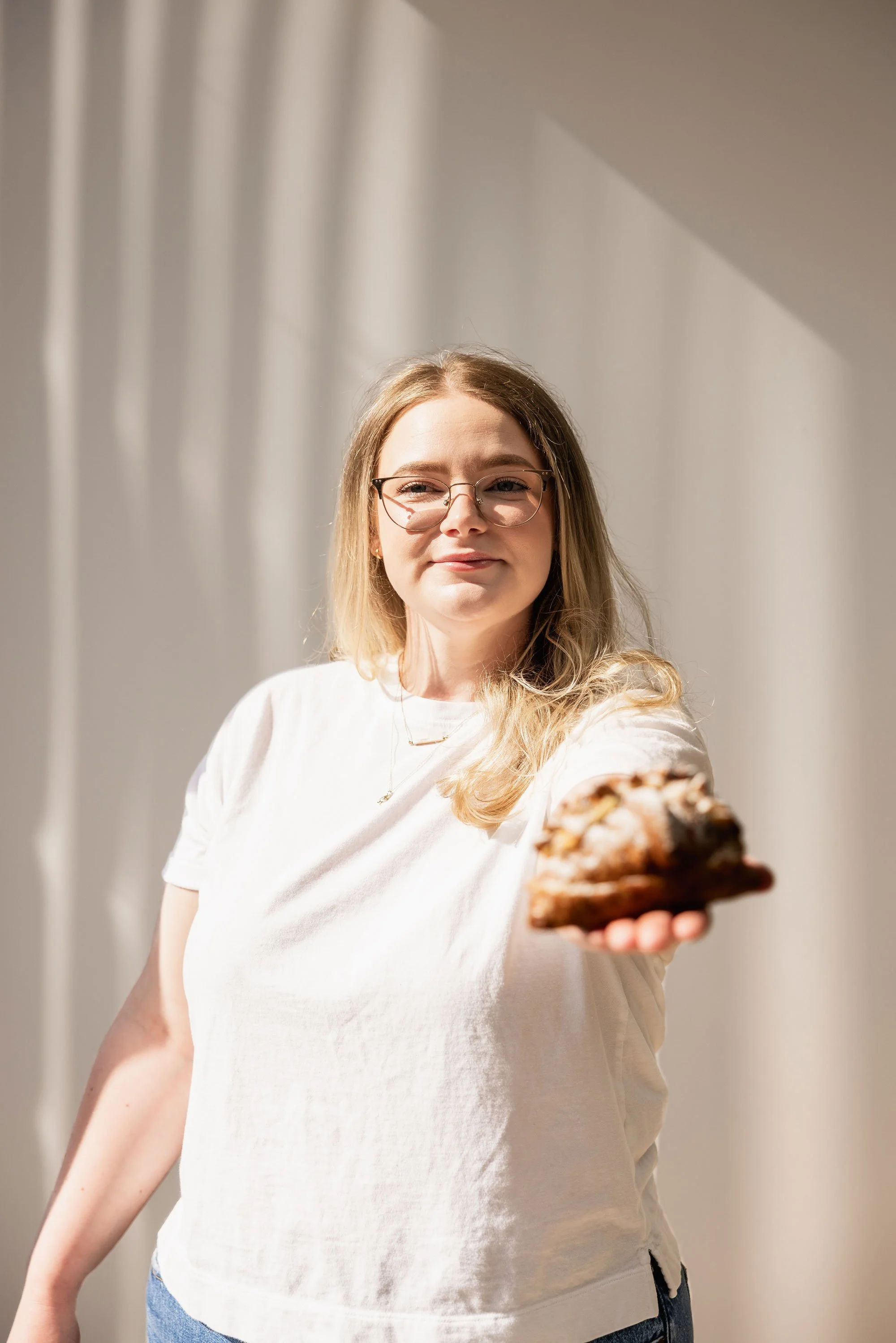 A woman with blonde hair, glasses, and a white t-shirt extends her hand forward, holding a piece of cake toward the camera indoors with white curtains in the background.