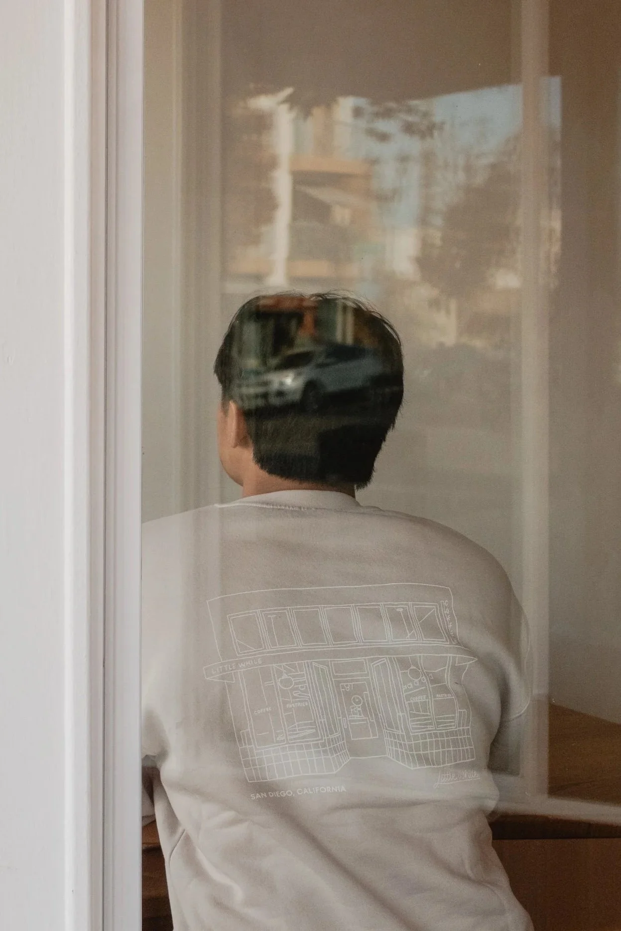 A person sitting with their back to the camera, seen through a glass reflection, with a city street scene visible on the glass.