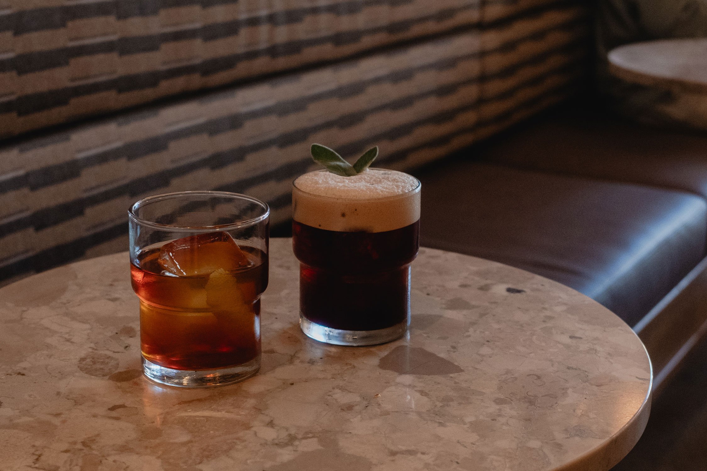 Two seasonal cocktail beverages on a marble table, one with ice and the other with foam and a small green garnish.